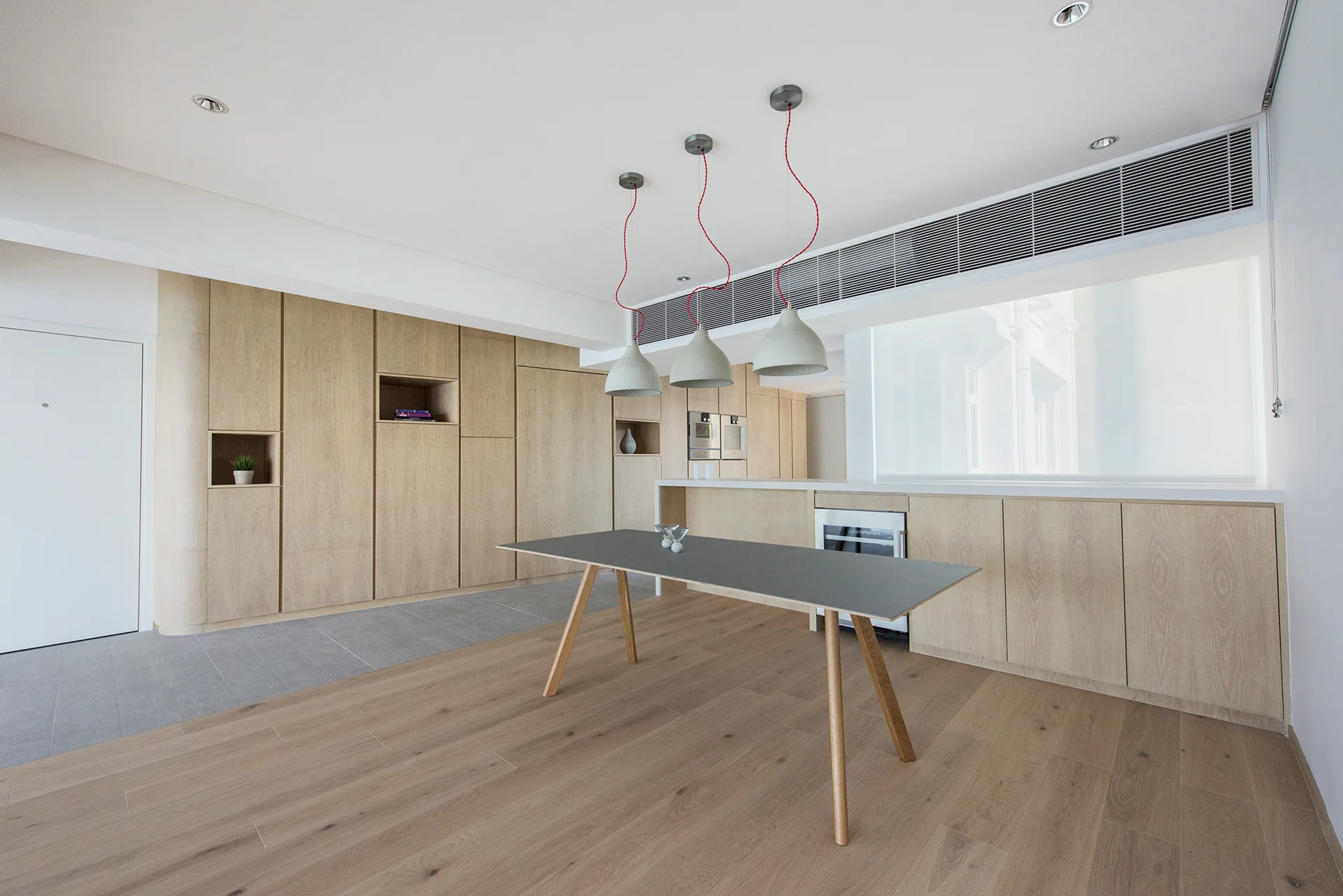 A minimalist kitchen features seamless light wood cabinetry, a grey dining table, and three dome pendant lights with bright red cords.