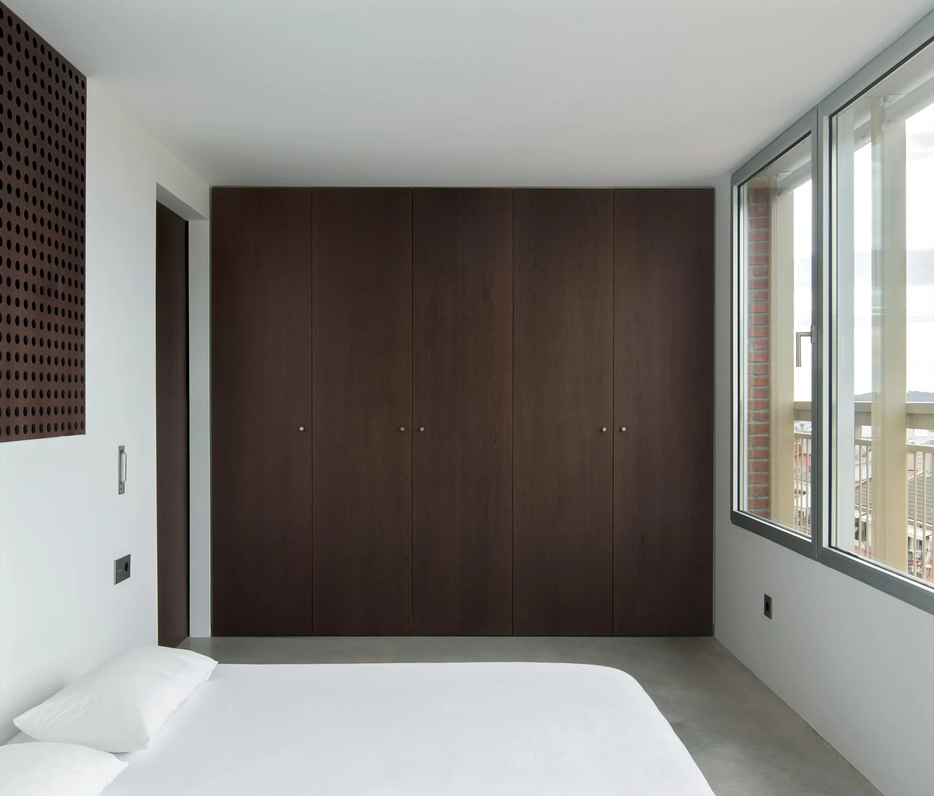 A minimalist bedroom featuring a large built-in dark wood wardrobe, plain white walls, a bed with white bedding, and large windows.