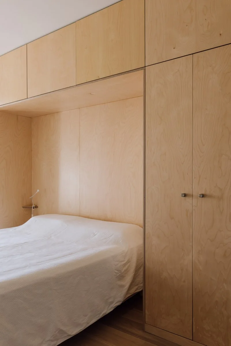 A minimalist bedroom features a bed tucked into a custom light plywood storage unit with flat-panel closets and overhead cabinets.