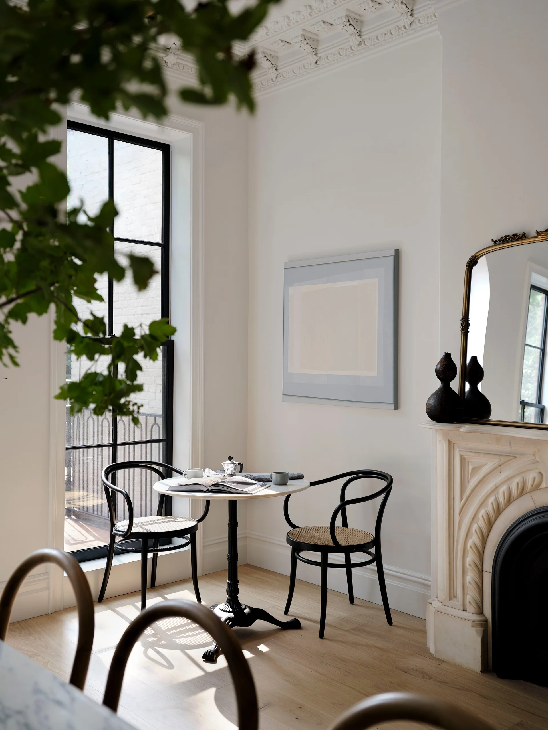 A small white bistro table and two black cane-seat chairs are set beside a tall black-framed window and a carved white marble fireplace.
