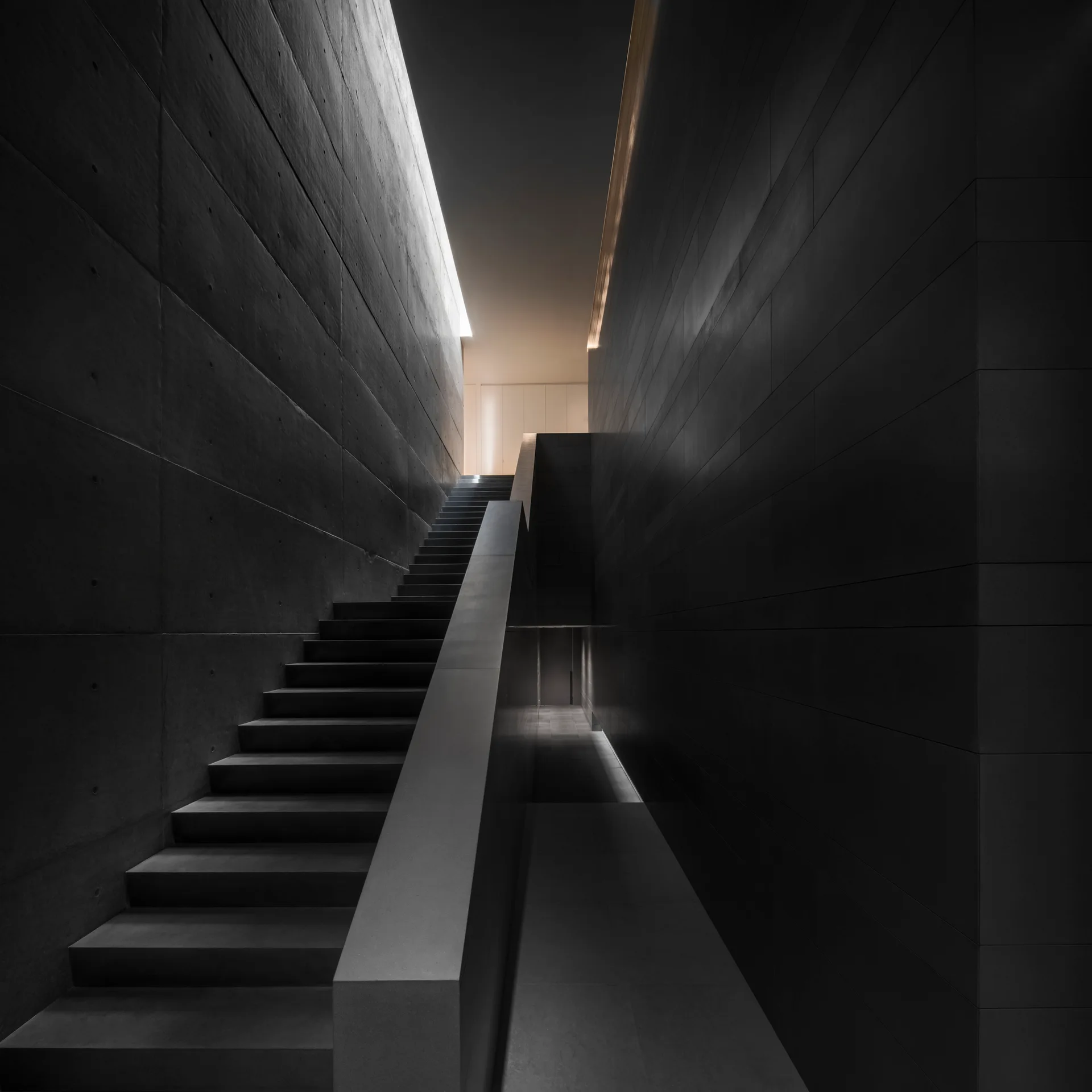 A dark gray concrete staircase rises between monolithic charcoal walls illuminated by indirect cove lighting along the ceiling.