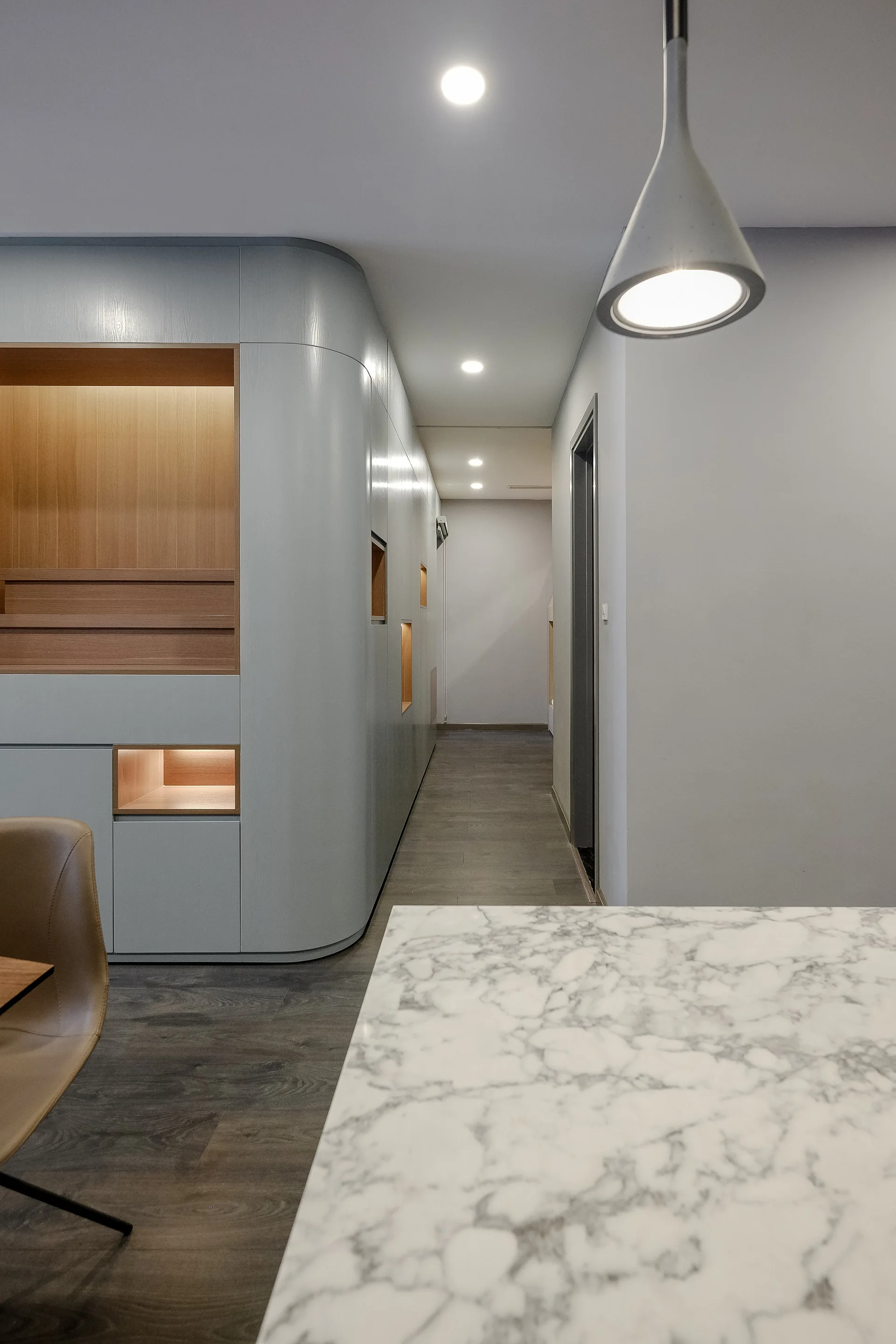 A modern hallway featuring a curved light-grey built-in wall unit with warm-toned illuminated wood niches next to a marble-topped island.