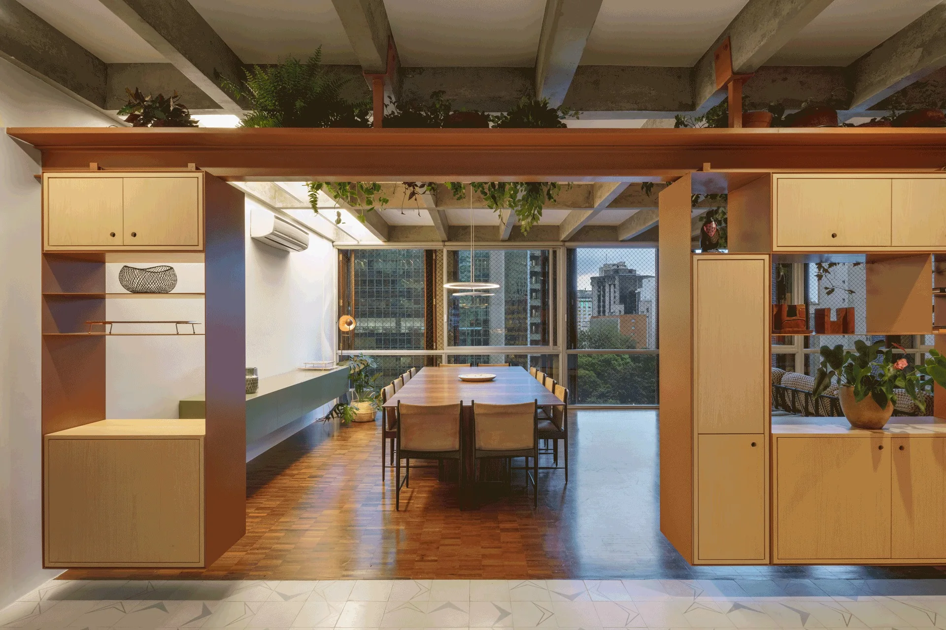 A long wooden dining table is flanked by custom storage dividers under an exposed concrete ceiling decorated with green plants.