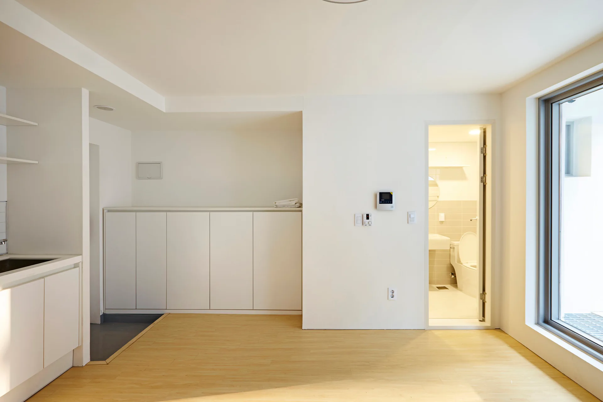 A minimalist room with light wood floors, plain white walls, a built-in white storage cabinet, and an open doorway leading to a tiled bathroom.