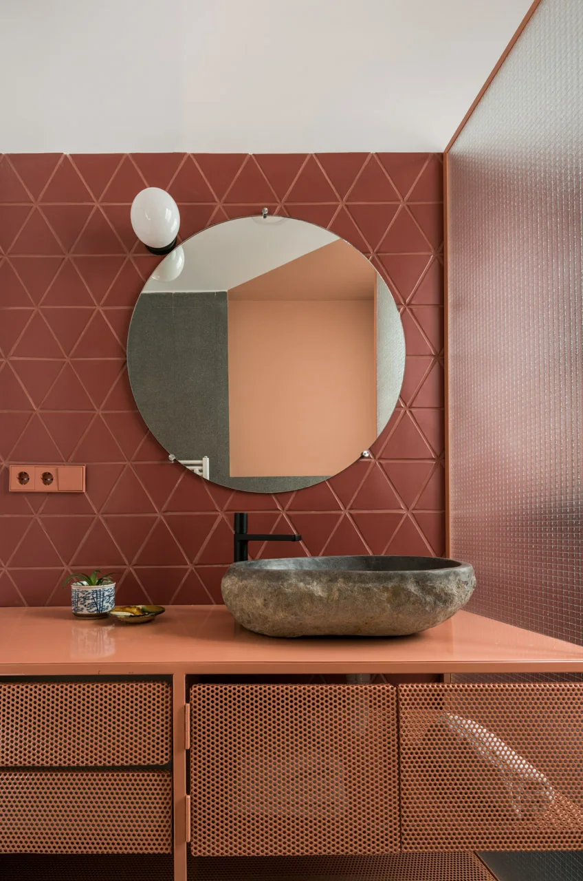 A modern bathroom vanity featuring a rustic stone vessel sink, terracotta triangular tiles, a round mirror, and a perforated orange cabinet.