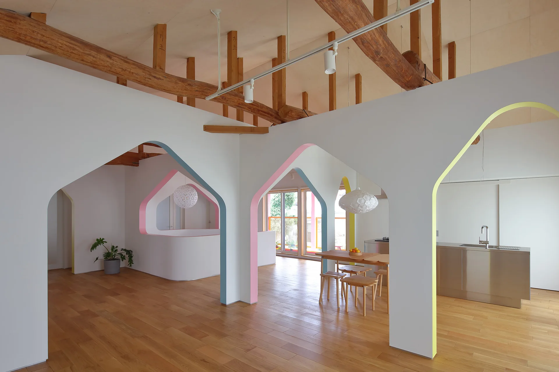 A minimalist open-plan interior featuring white partition walls with pastel-trimmed archways, light wood flooring, and exposed ceiling beams.