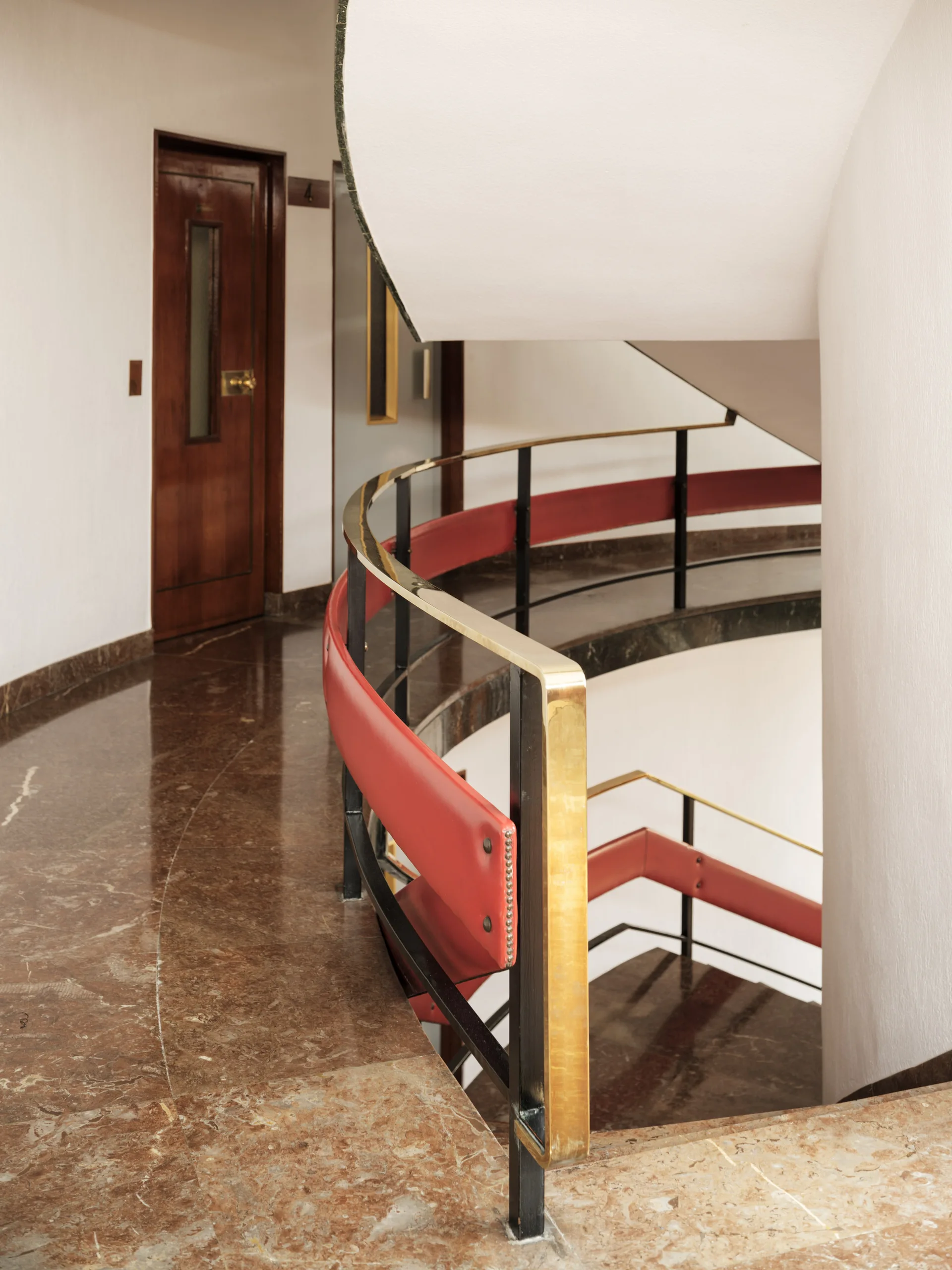 A curved staircase features a polished brass and red leather handrail alongside brown marble floors and white walls.