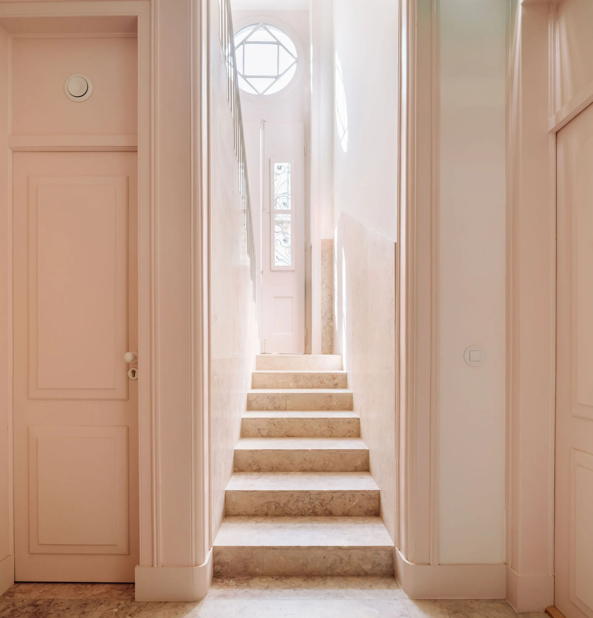 A narrow hallway features a stone staircase leading up to a round window, flanked by blush pink paneled doors and trim.