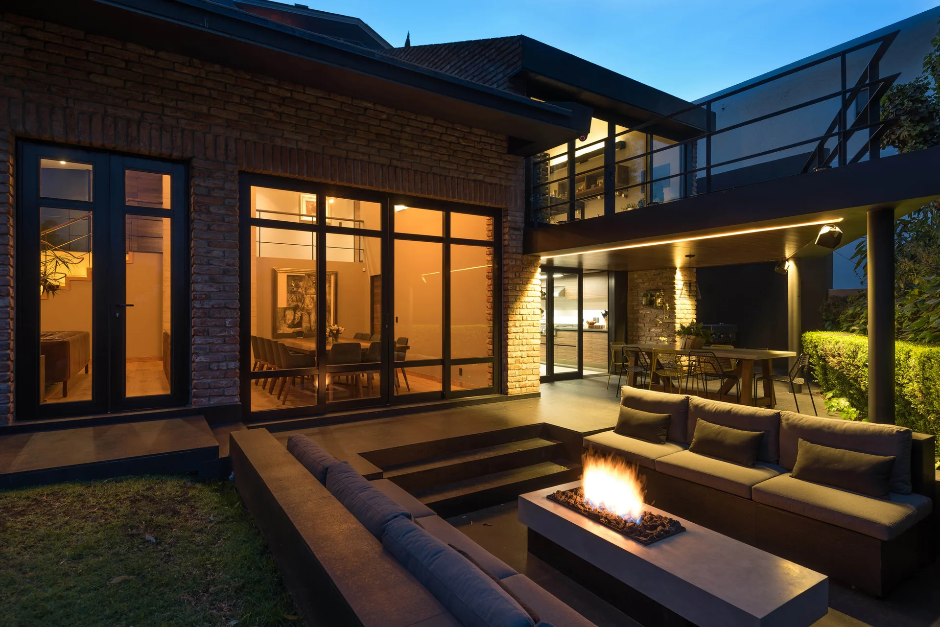 A modern backyard patio features a sunken lounge with a glowing fire pit next to an exposed brick home with large windows.