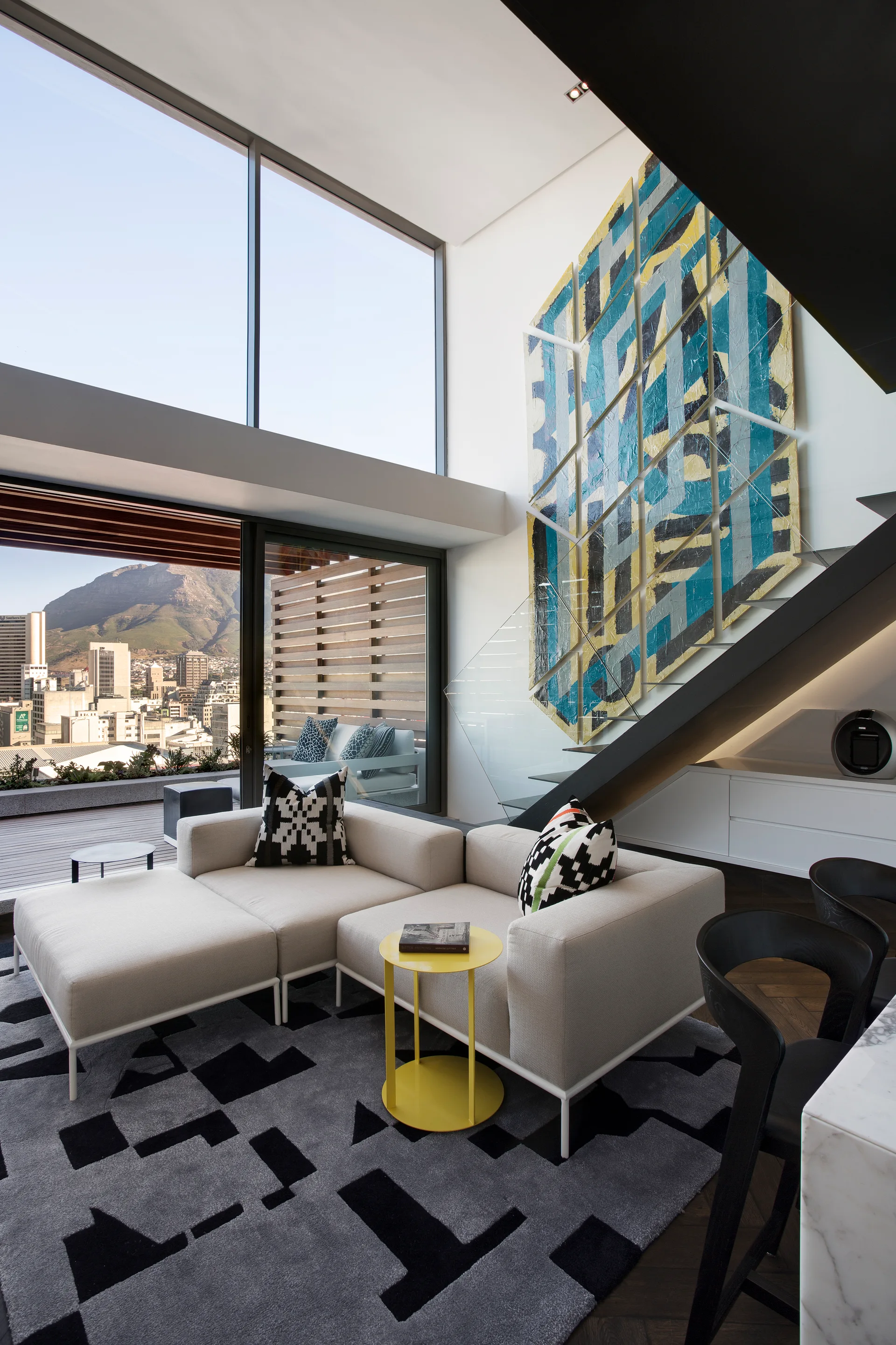 A high-ceilinged living room features a white modular sectional, a large geometric painting beside a glass staircase, and a bold patterned rug.