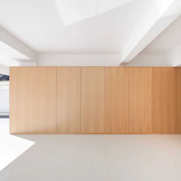 A seamless wall of floor to ceiling light wood cabinetry sits between a smooth white floor and a white beamed ceiling in a minimalist room.