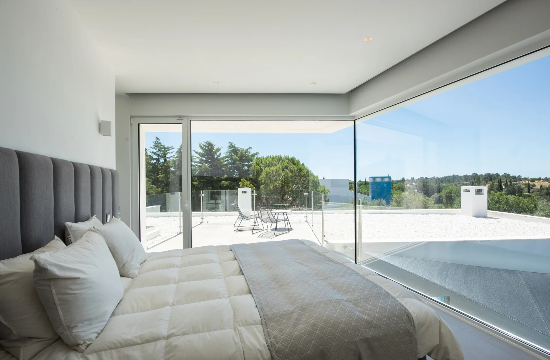 A minimalist bedroom featuring a grey channel-tufted headboard, white bedding, and floor-to-ceiling glass walls that look out onto a terrace.