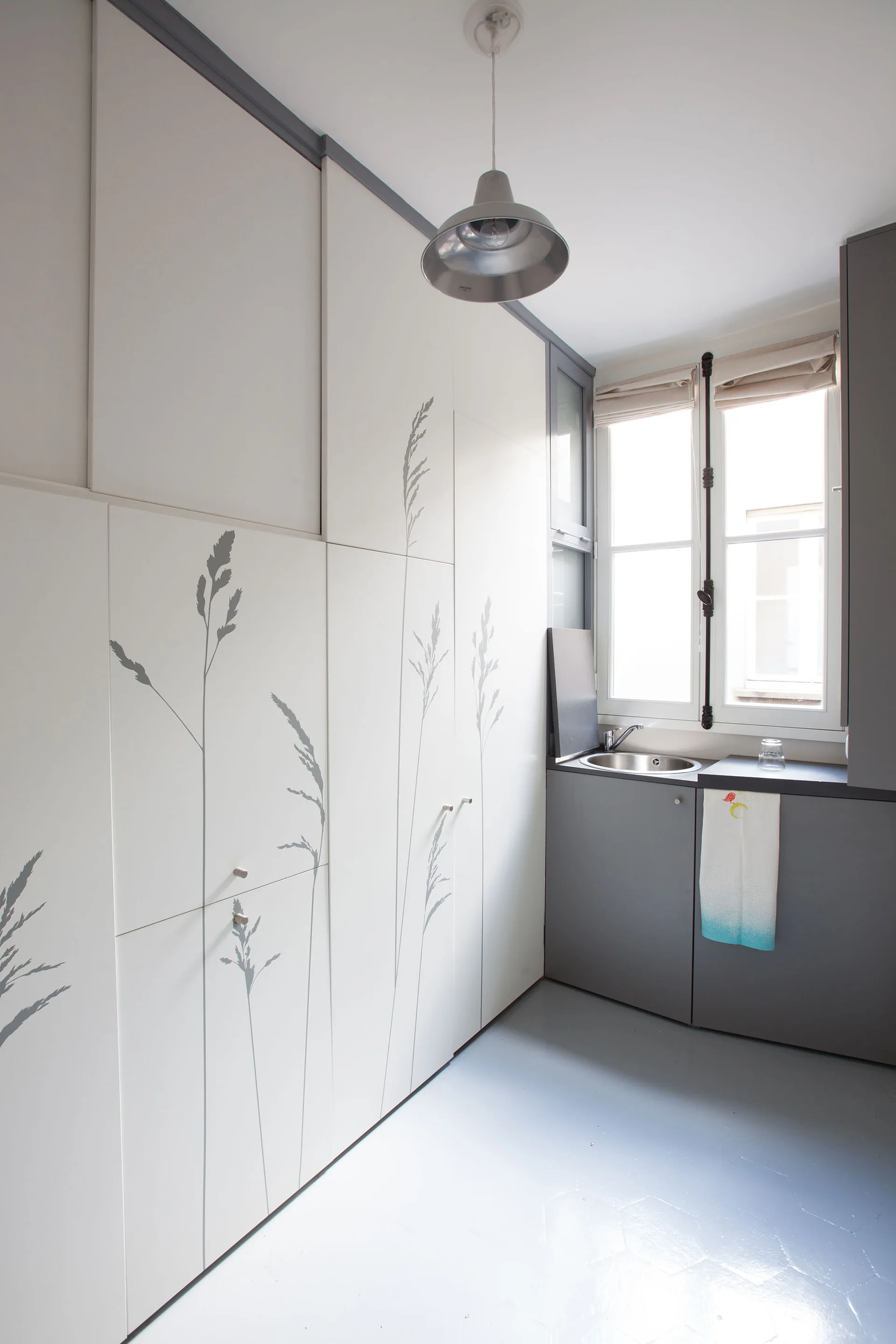 A compact modern kitchen with tall white storage cabinets featuring gray botanical illustrations, a small gray counter, and a metal pendant light.