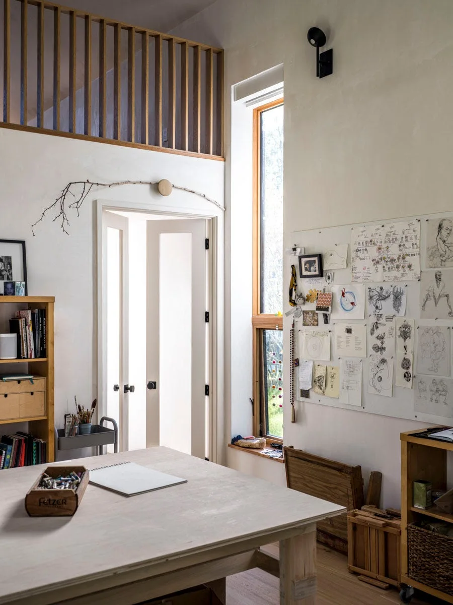 A bright creative studio space featuring a large wooden desk, a tall narrow window, a white door, and a bulletin board covered with sketches.