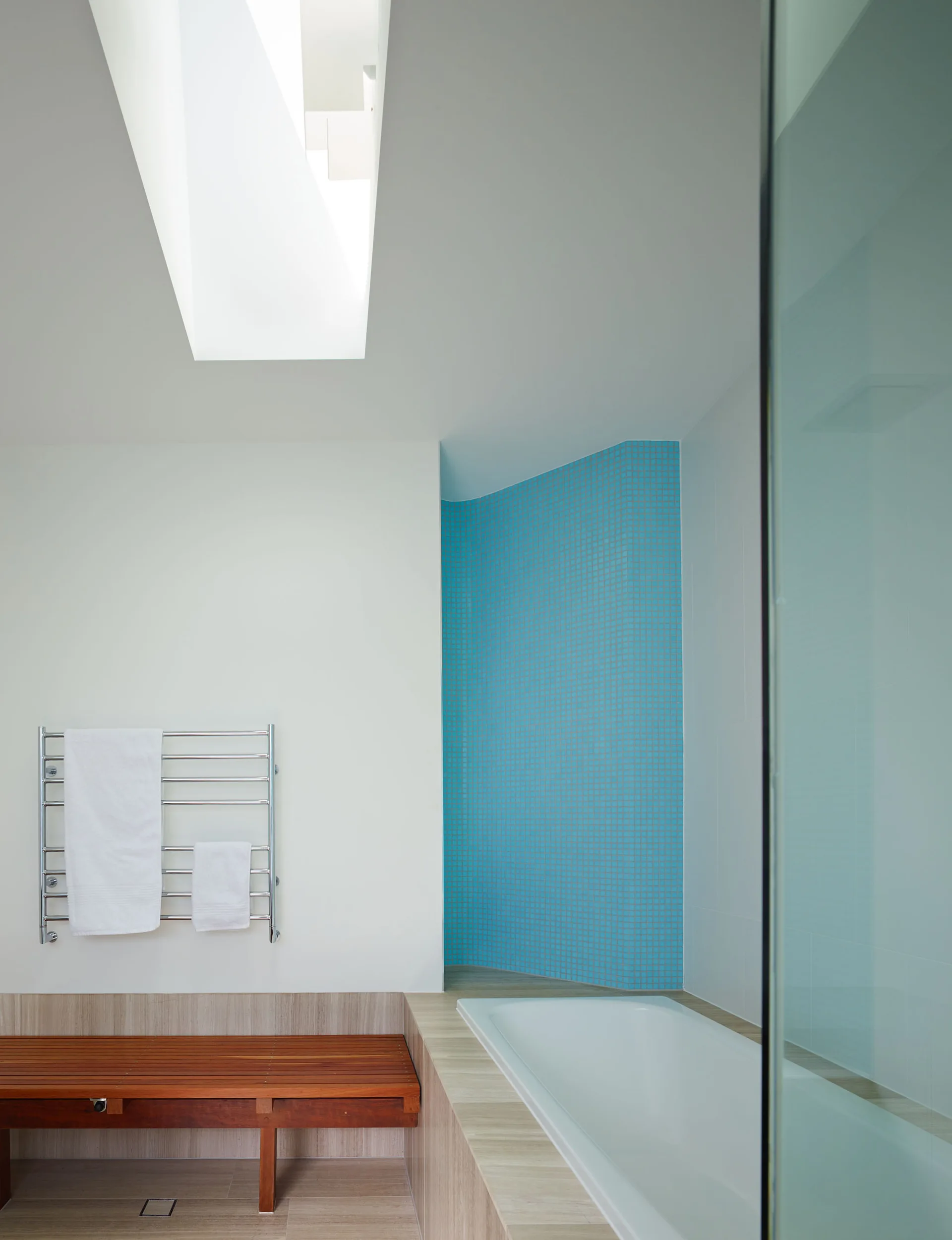A modern bathroom with a white bathtub, a curved blue mosaic tile wall, a slatted wood bench, and a skylight in the white ceiling.
