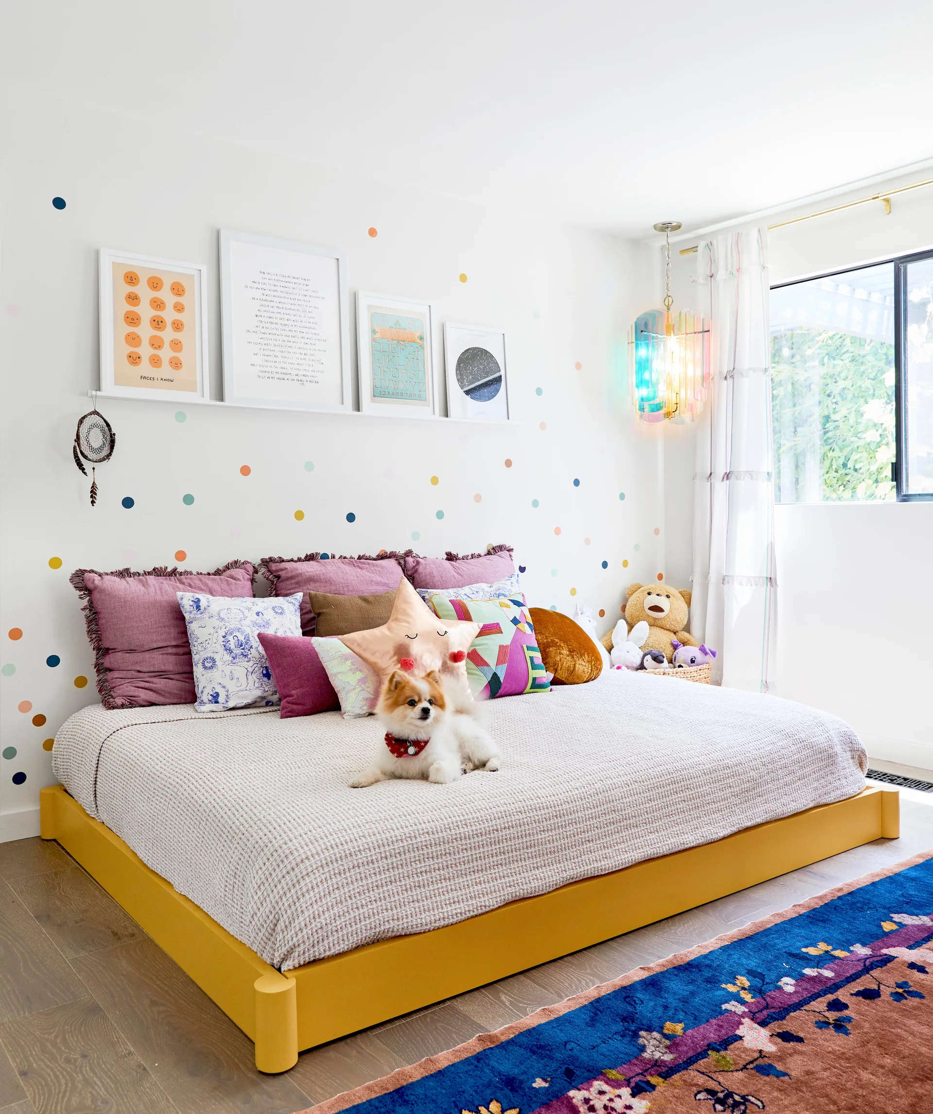 A kids bedroom features a yellow platform bed, a wall decorated with colorful polka dots, framed art on a display shelf, and an iridescent pendant.