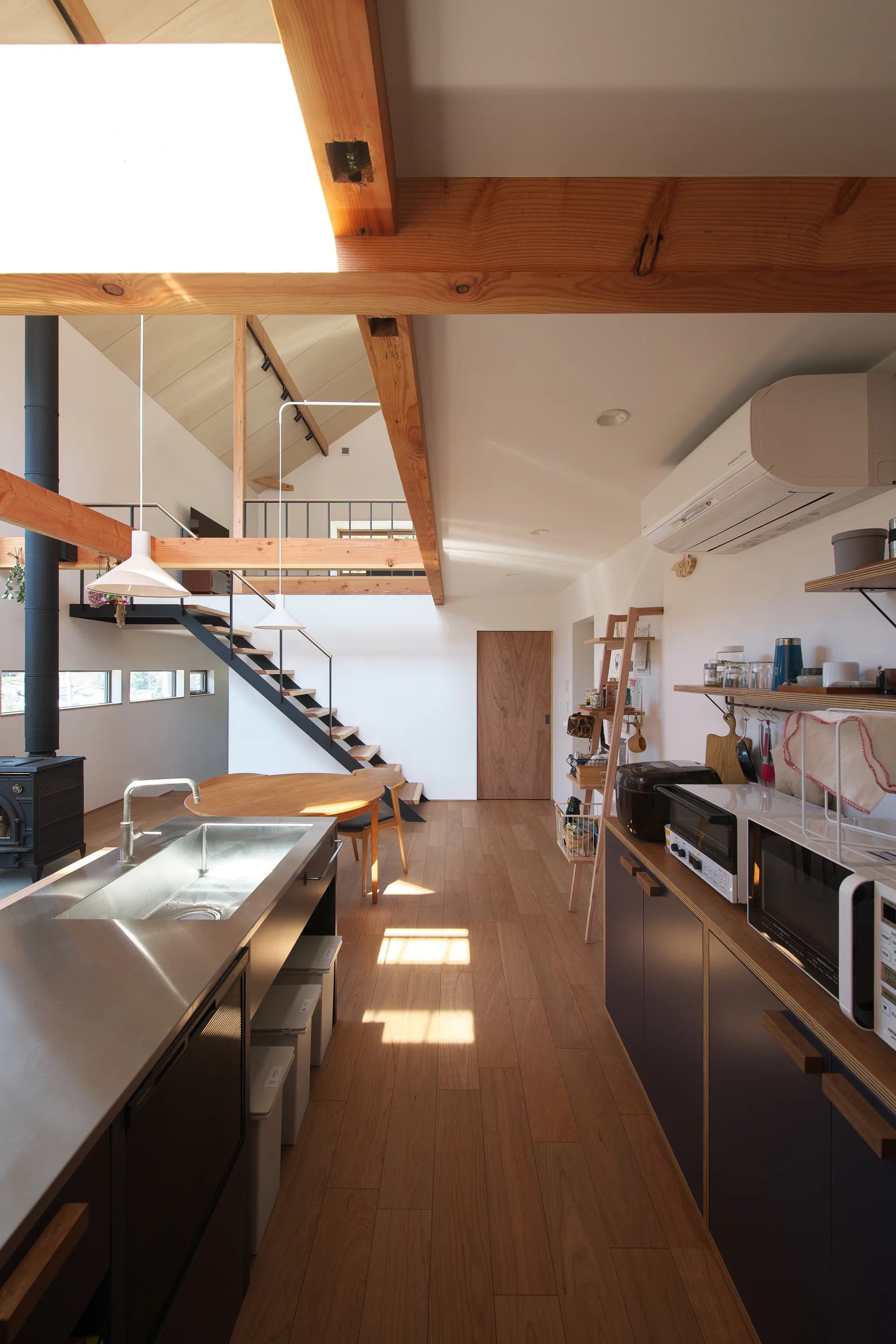 An open-plan kitchen features a stainless steel island, dark grey cabinets, wood floors, and exposed timber ceiling beams beneath a mezzanine loft.