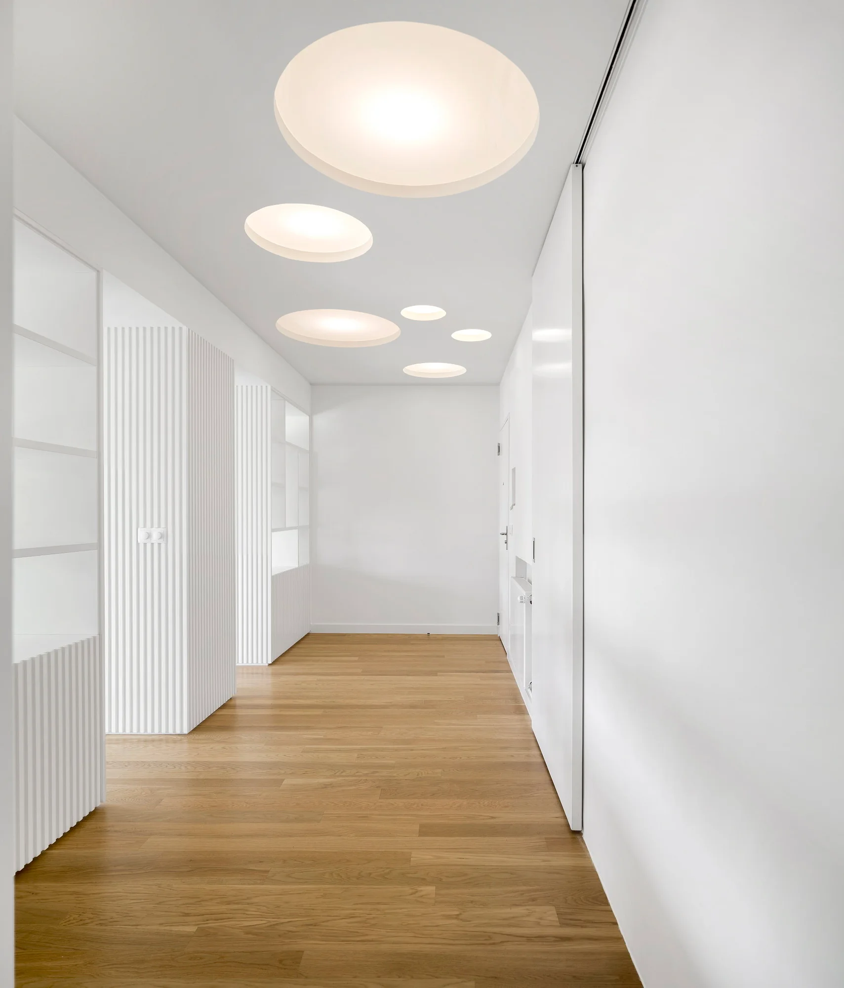 A minimalist white hallway featuring light wood floors, circular recessed ceiling lights, and built-in shelving with vertical fluted paneling.