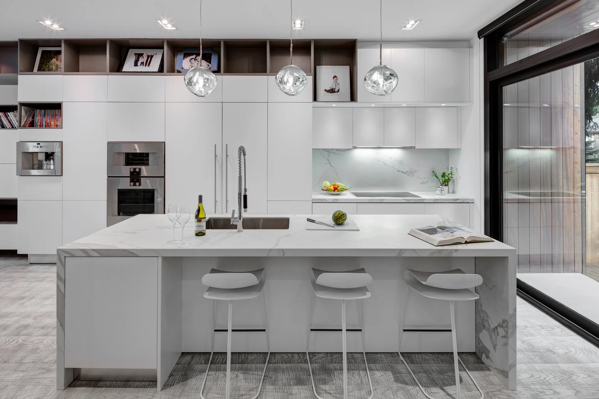 A modern white kitchen features a marble waterfall island with bar stools, handleless flat-panel cabinets, and three hanging glass globe pendant lights.