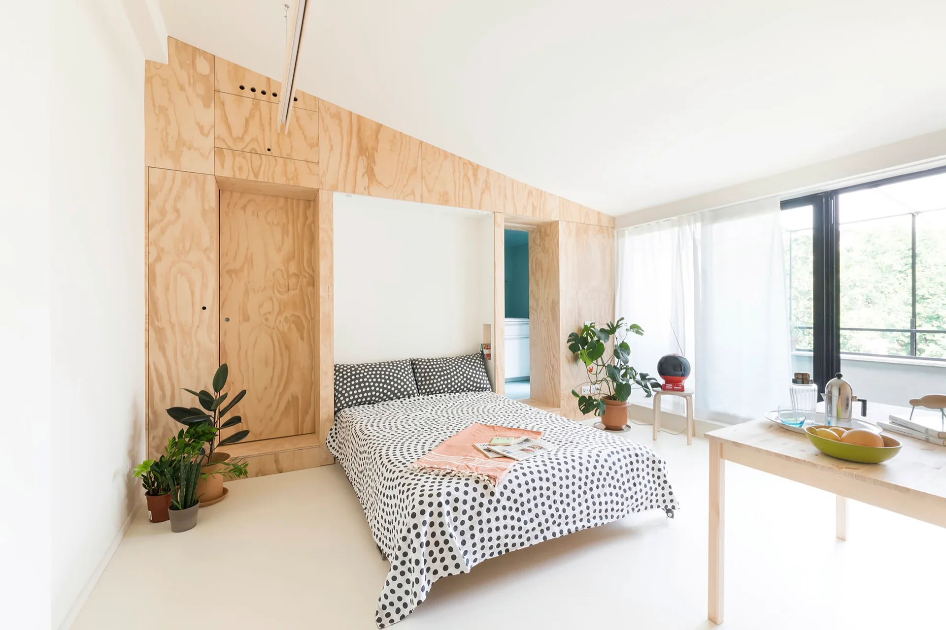 A minimalist bedroom features a bed with polka dot sheets framed by a large light plywood built-in wall unit under a sloped ceiling.