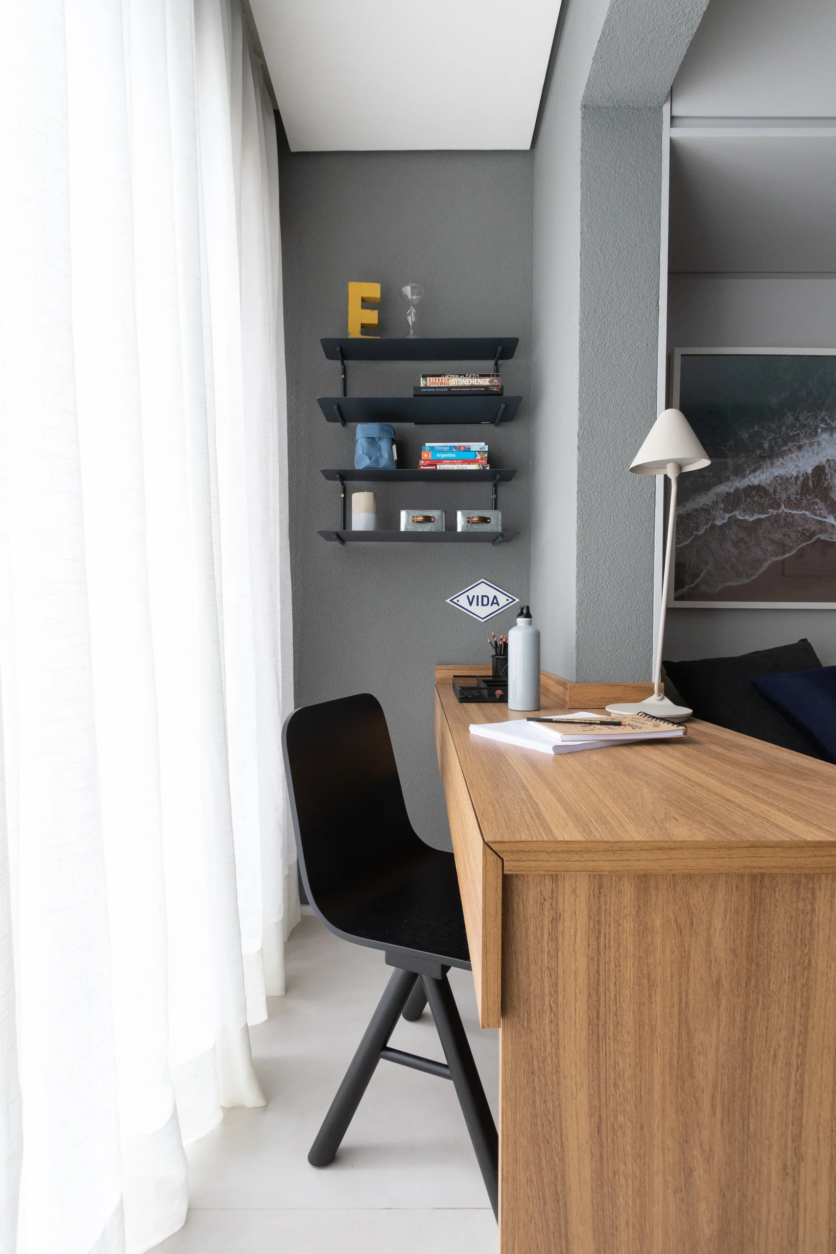 A minimalist home office features a wood desk, a black swivel chair, and small black floating shelves on a textured grey wall by sheer curtains.