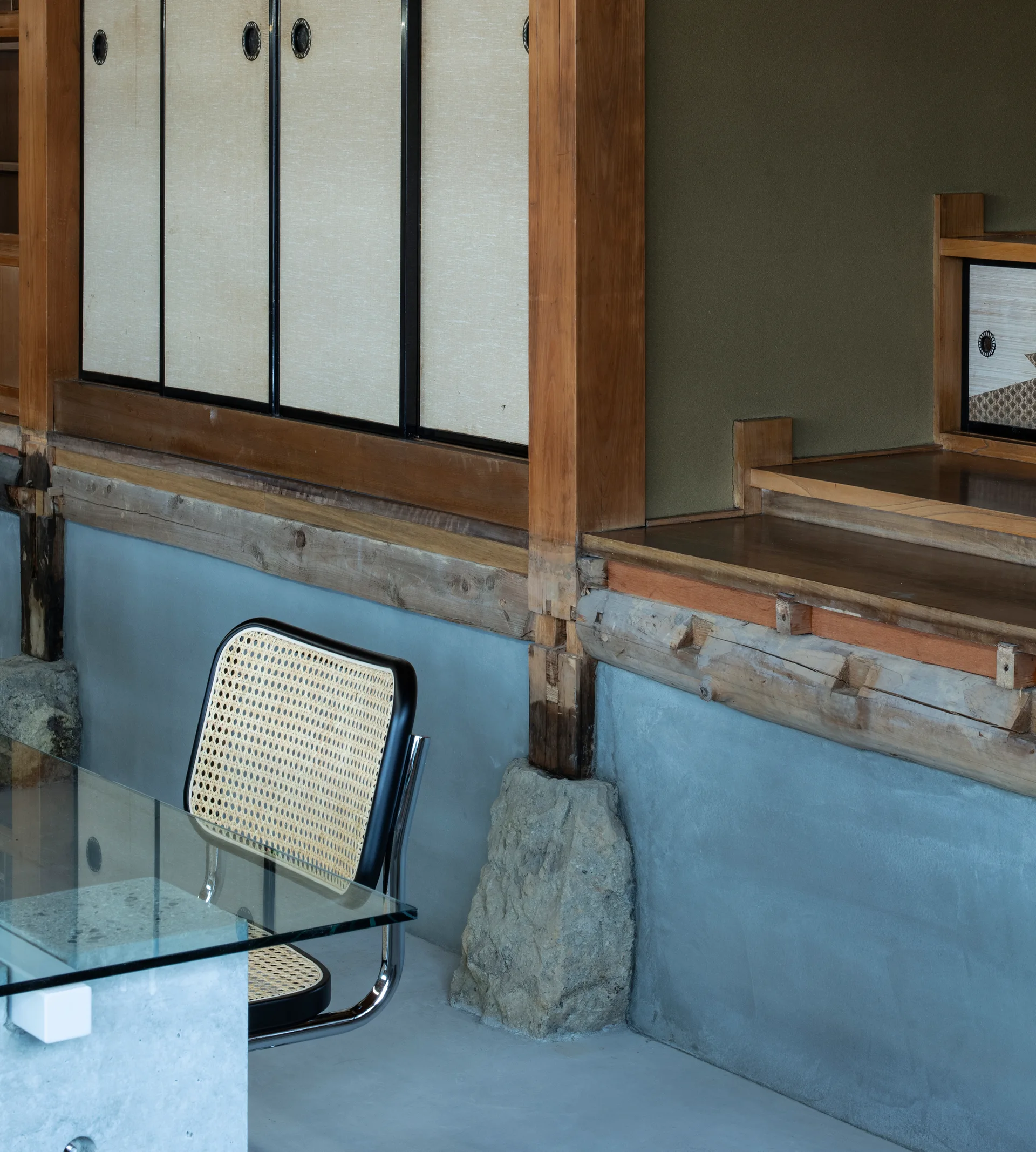 A cane-backed chair and a glass-top desk sit in a minimalist room with traditional Japanese sliding doors and exposed wooden framing.