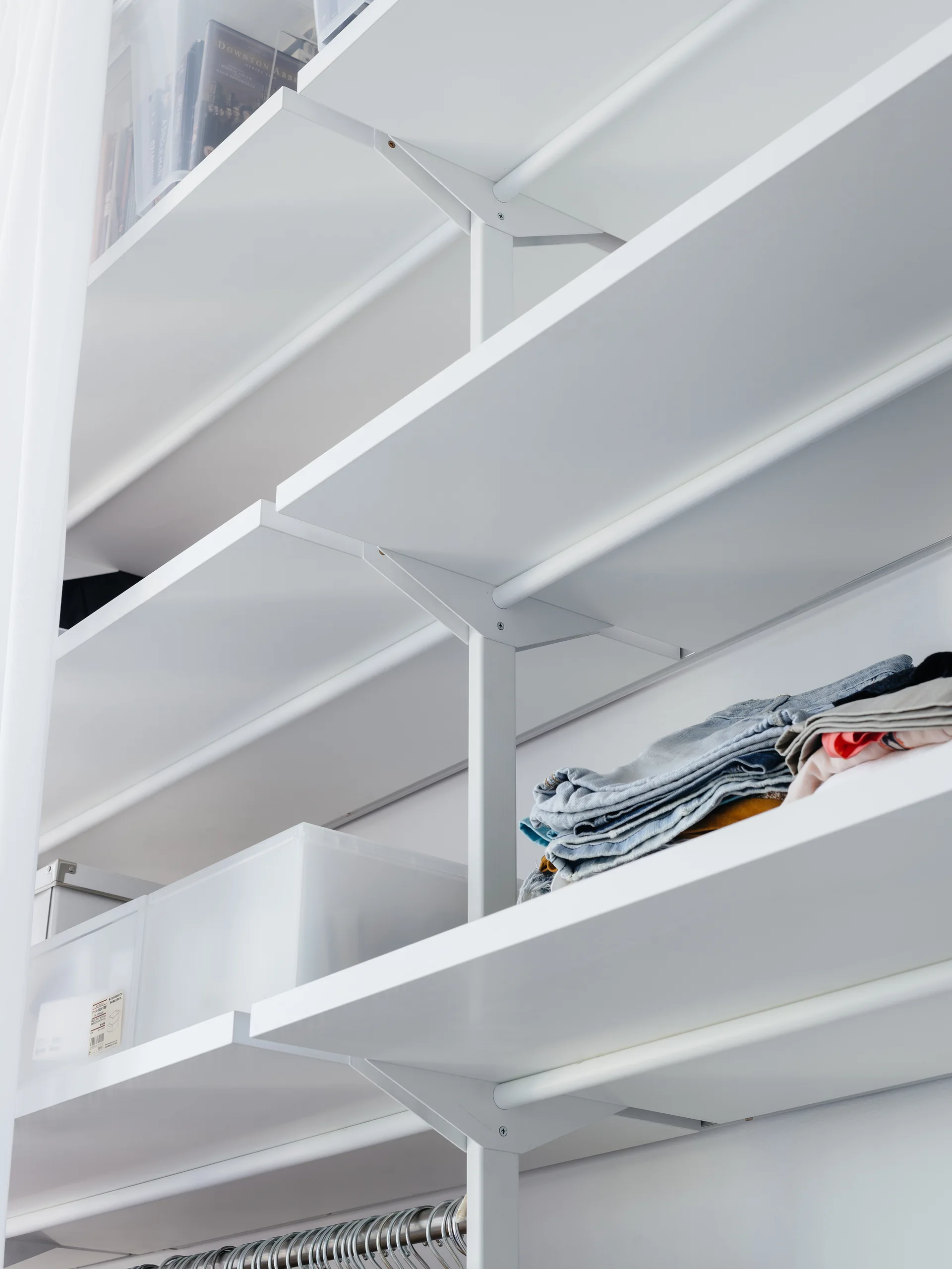 A close-up view of a white modular closet shelving system holding folded jeans and translucent plastic storage bins against a white wall.
