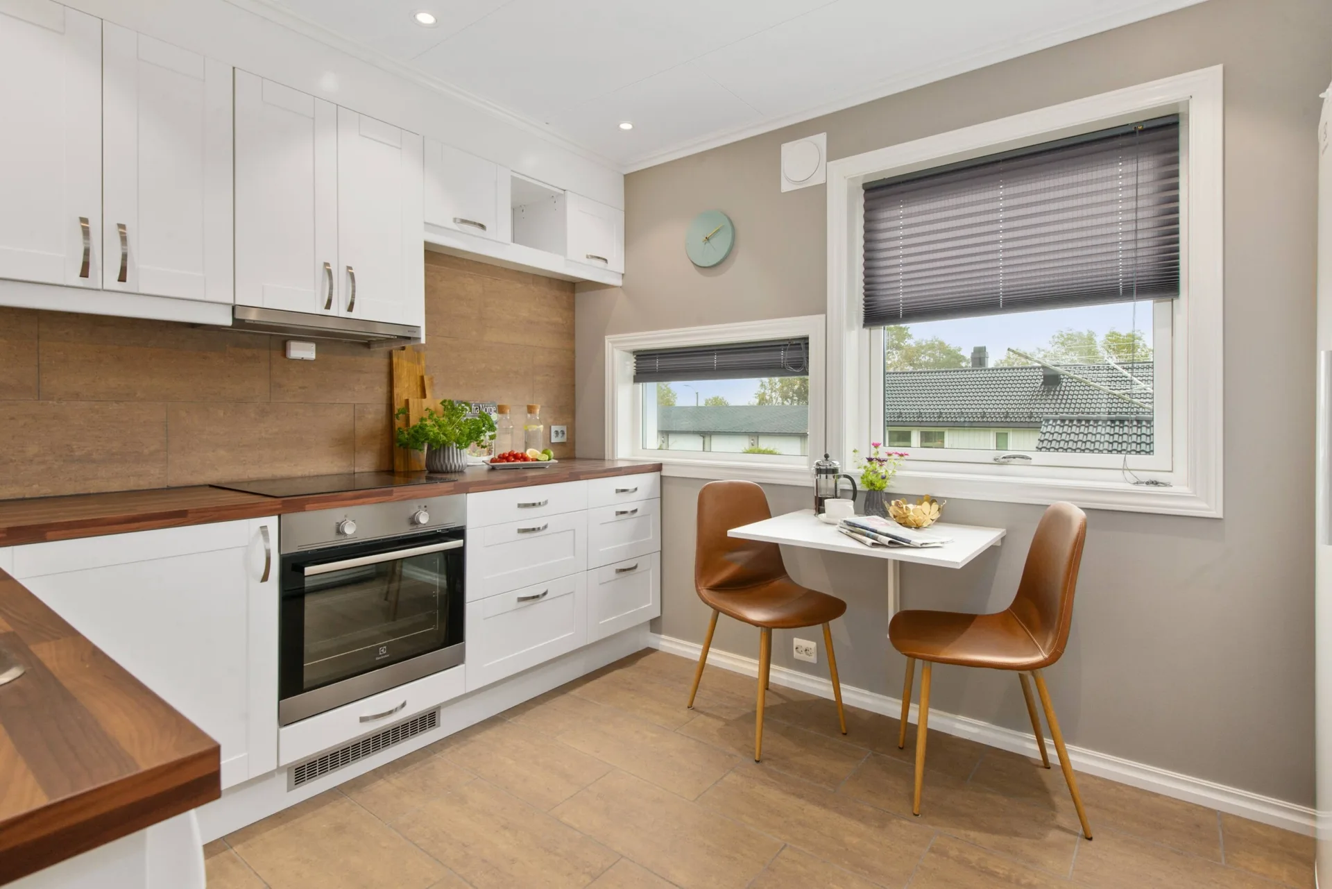 A compact Scandinavian kitchen features white cabinetry, a wood-toned backsplash, and a wall-mounted dining table with two brown chairs.