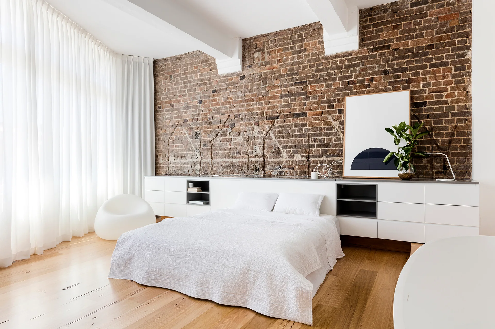 A minimalist bedroom features a low white bed, an exposed brick wall, a long white storage console, and light wood flooring.