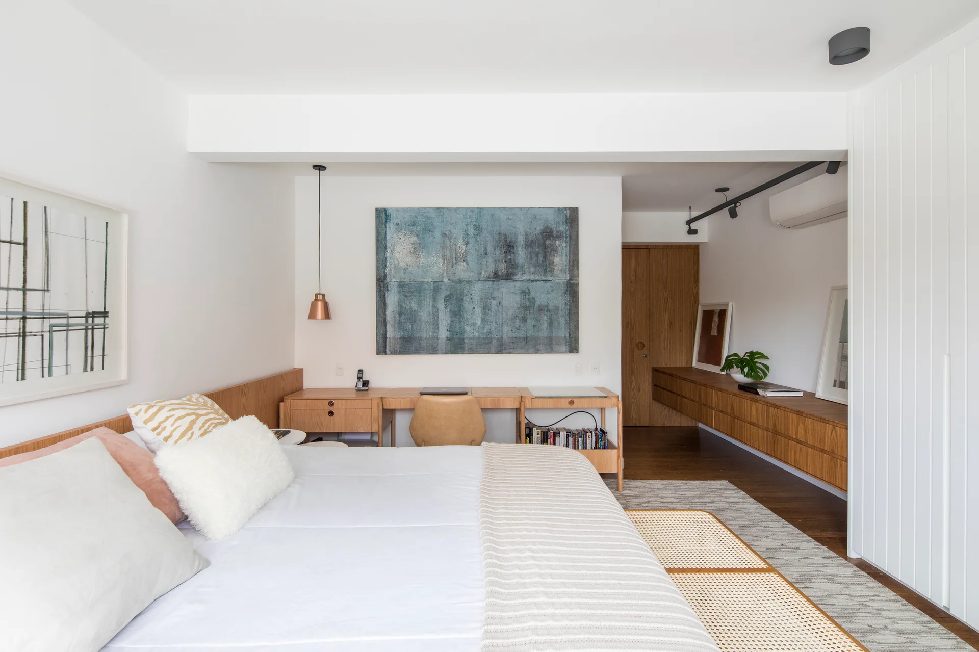 A modern bedroom features a bed with neutral linens, an integrated wooden headboard and desk unit, and a long floating cabinet.