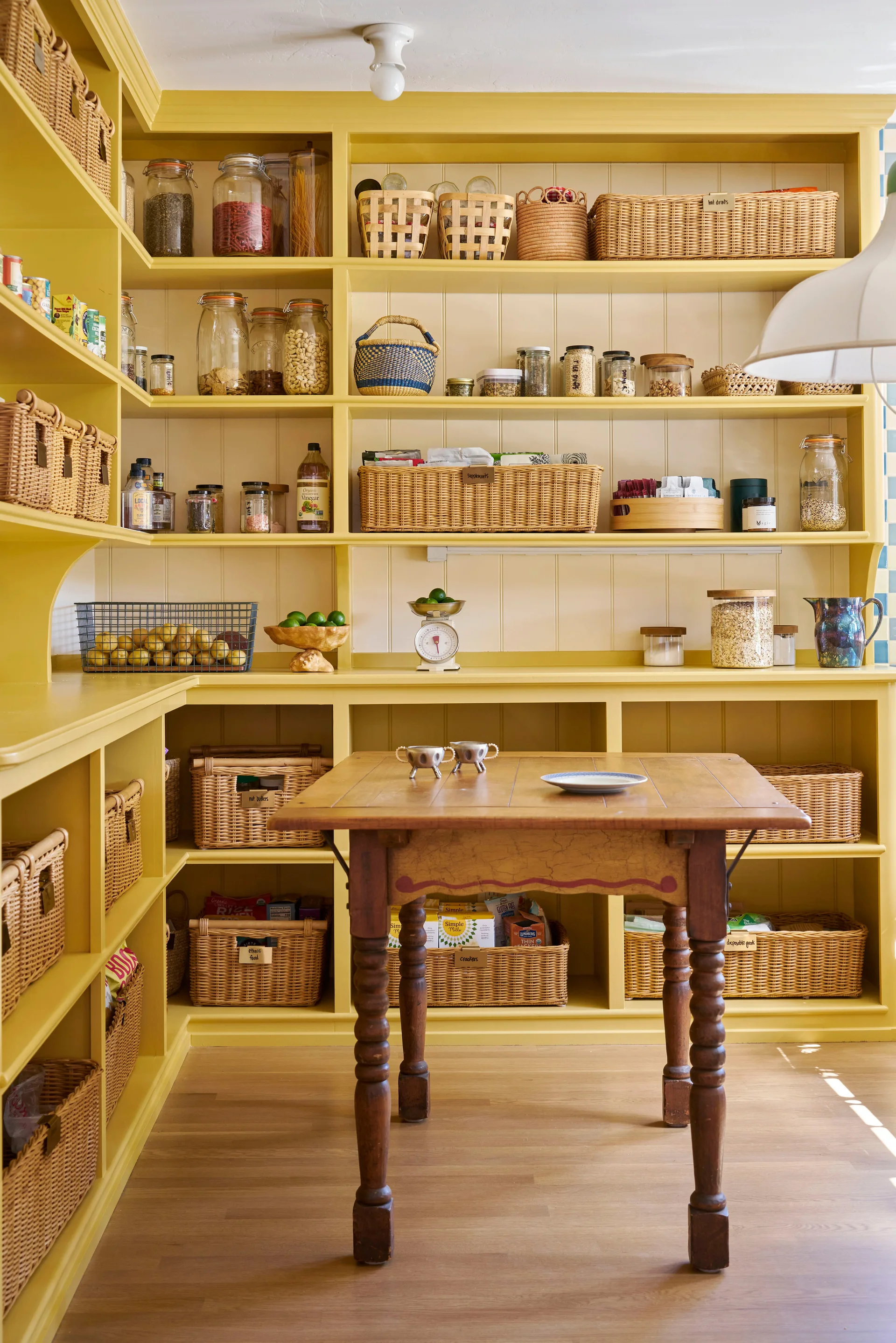 A corner pantry with bright yellow open shelving filled with wicker baskets and glass jars around a central antique wooden table.