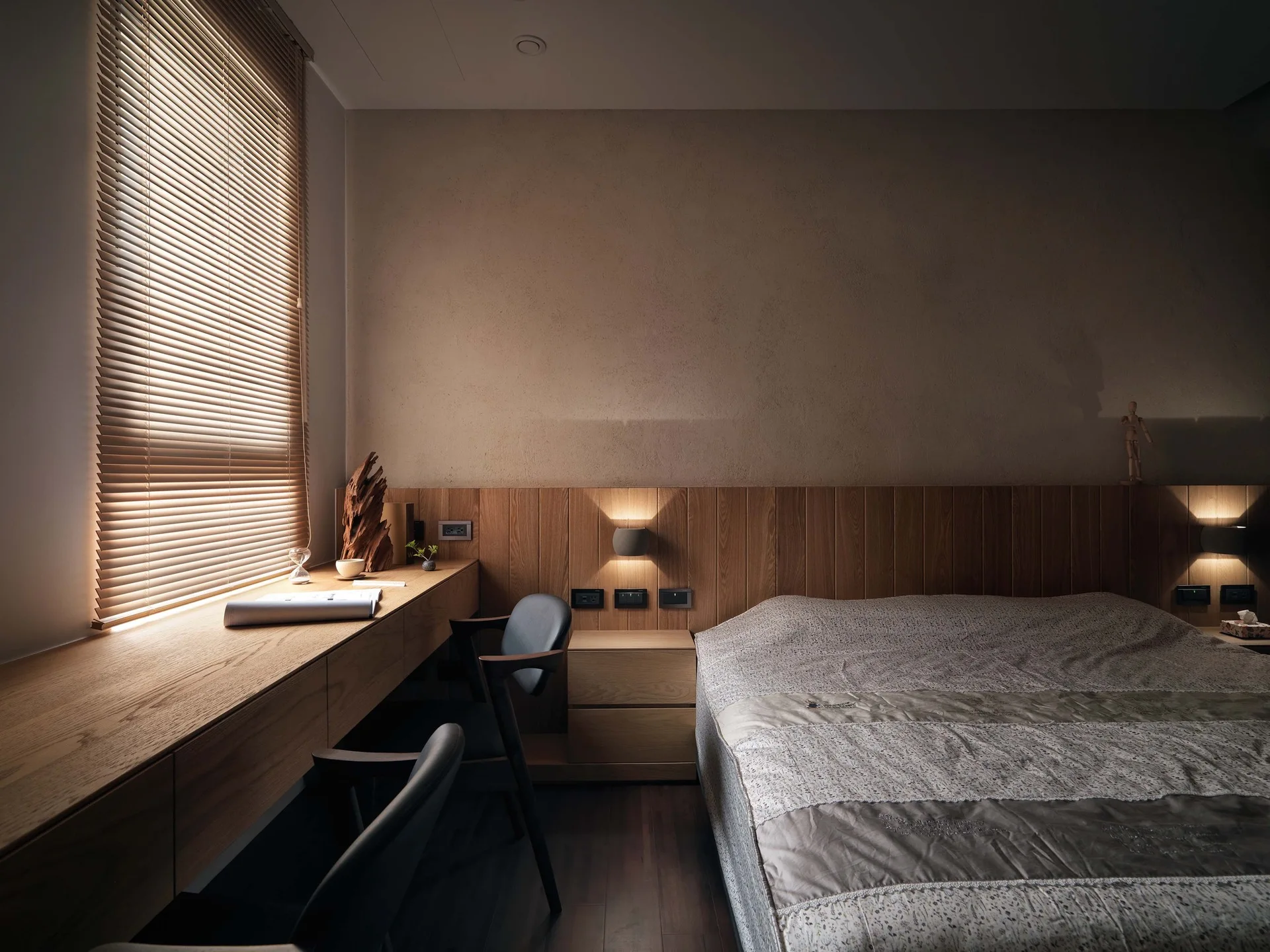 A minimalist bedroom features a long built-in wooden desk under a window with blinds, adjacent to a bed with a wood-slat headboard.
