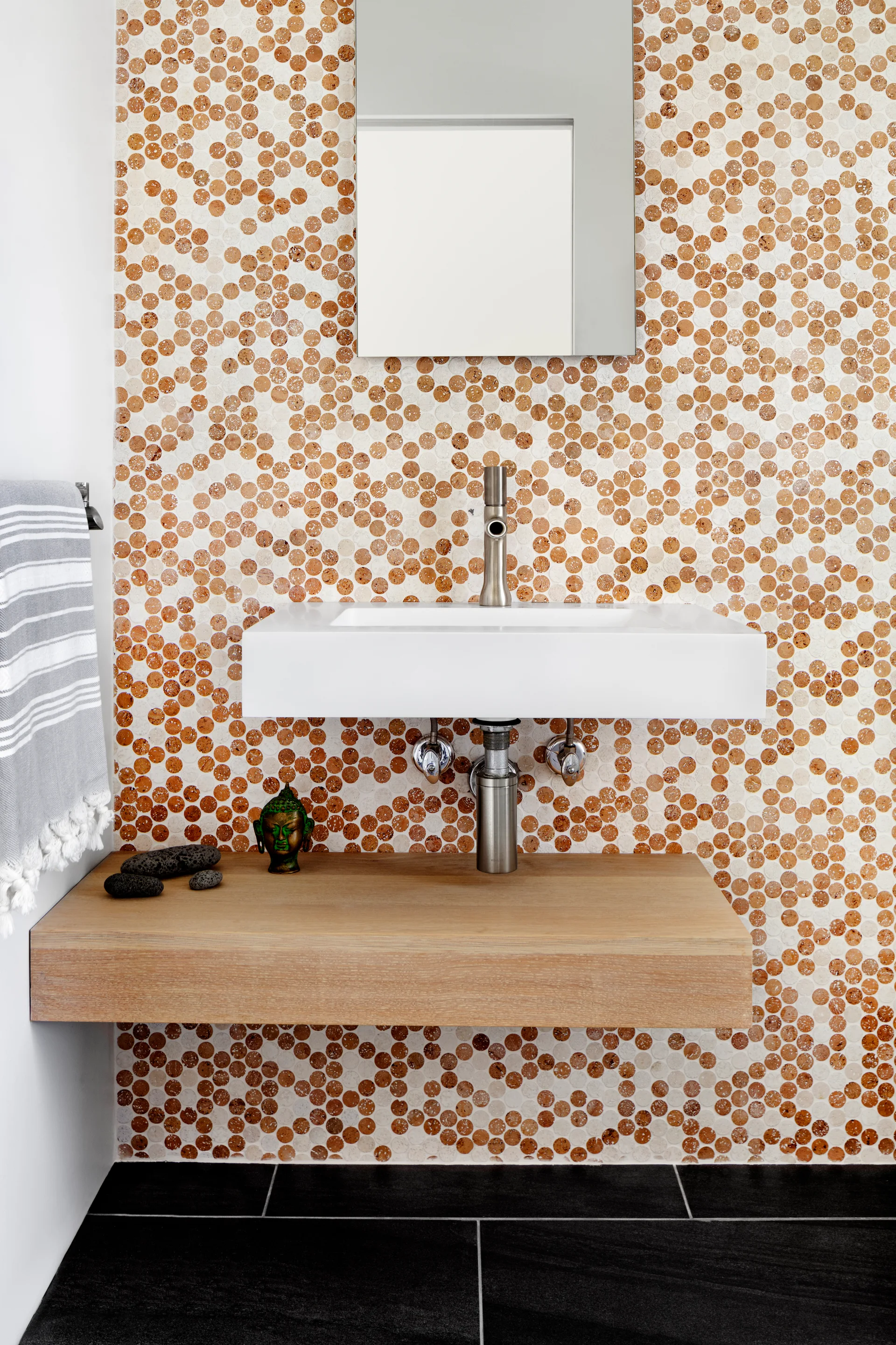A modern bathroom features a wall-mounted white sink and floating wood shelf against a patterned brown and beige penny tile wall.