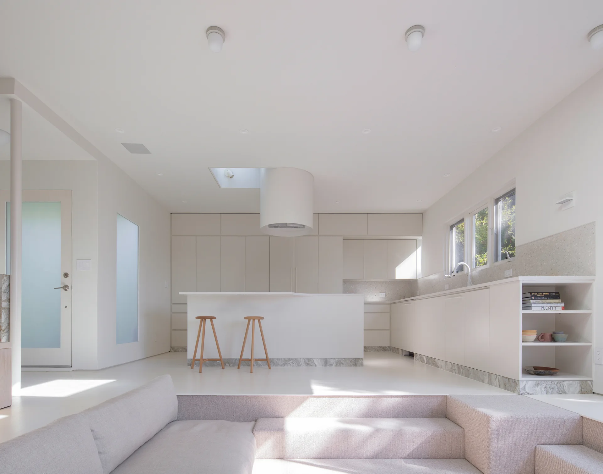 A minimalist beige kitchen with flat-front cabinetry and an island, stepping down into a sunken living area with a light-colored sofa.