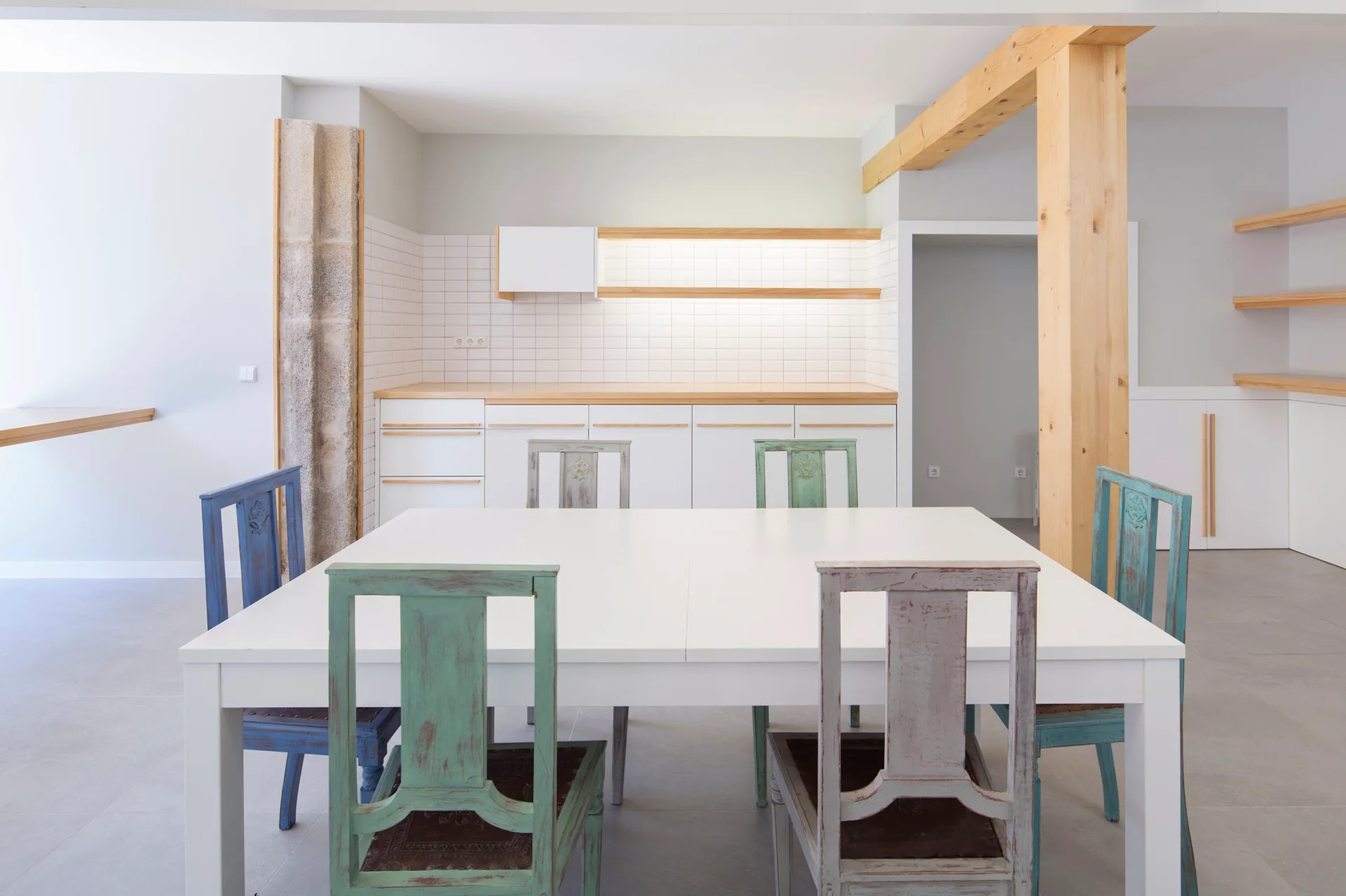 A white rectangular dining table surrounded by distressed green and blue wooden chairs in front of a white minimalist kitchen.