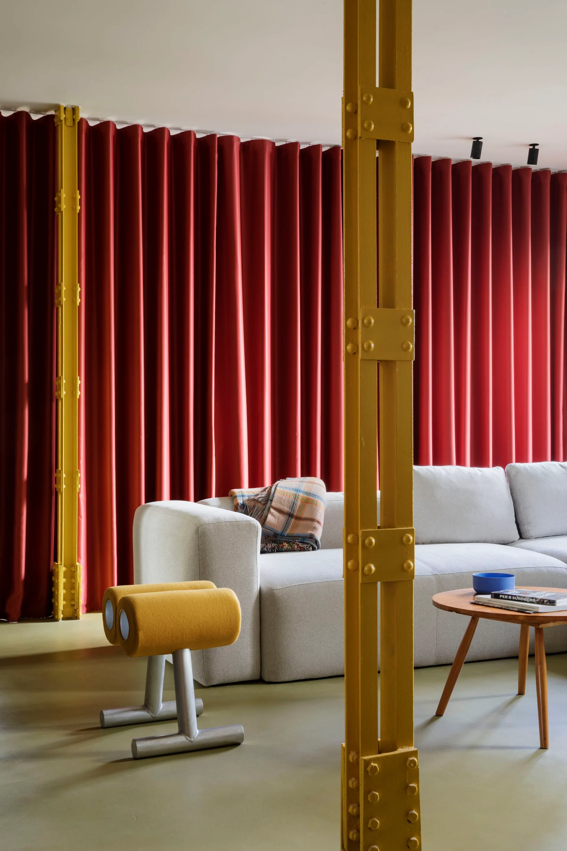 A modern grey sectional sofa sits in front of a full wall of pleated red curtains, framed by bright yellow painted industrial steel columns.
