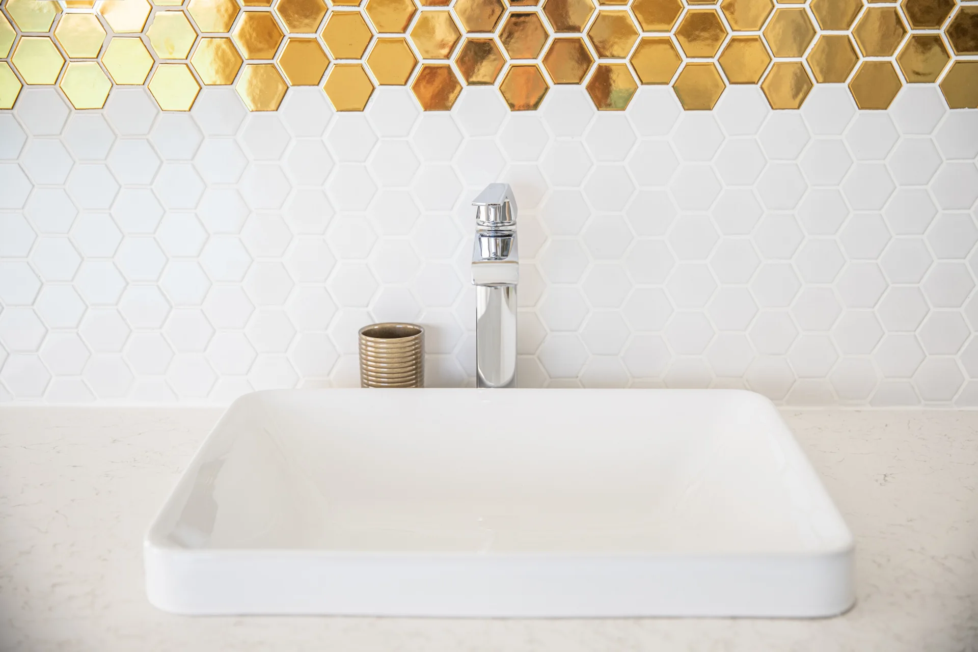 A close-up of a white vessel sink on a vanity with a white and gold hexagonal tile backsplash.