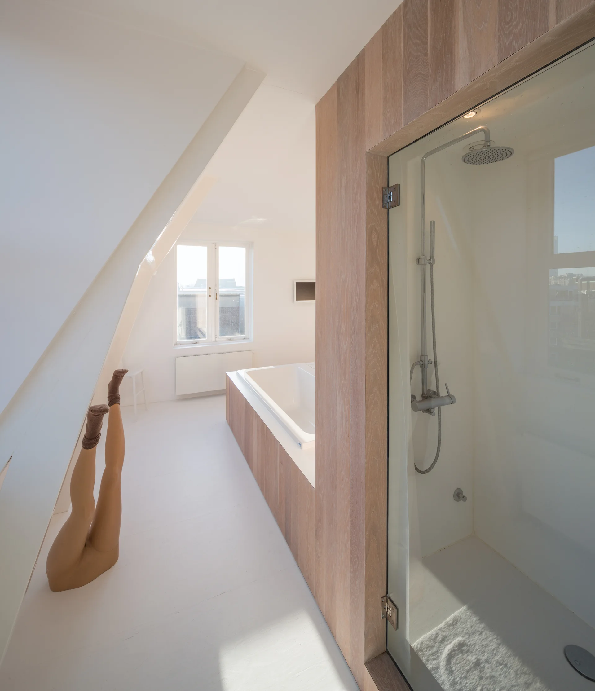 A minimalist attic bathroom features white floors, a sloped ceiling, and a built-in bathtub clad in light wood next to a glass shower.