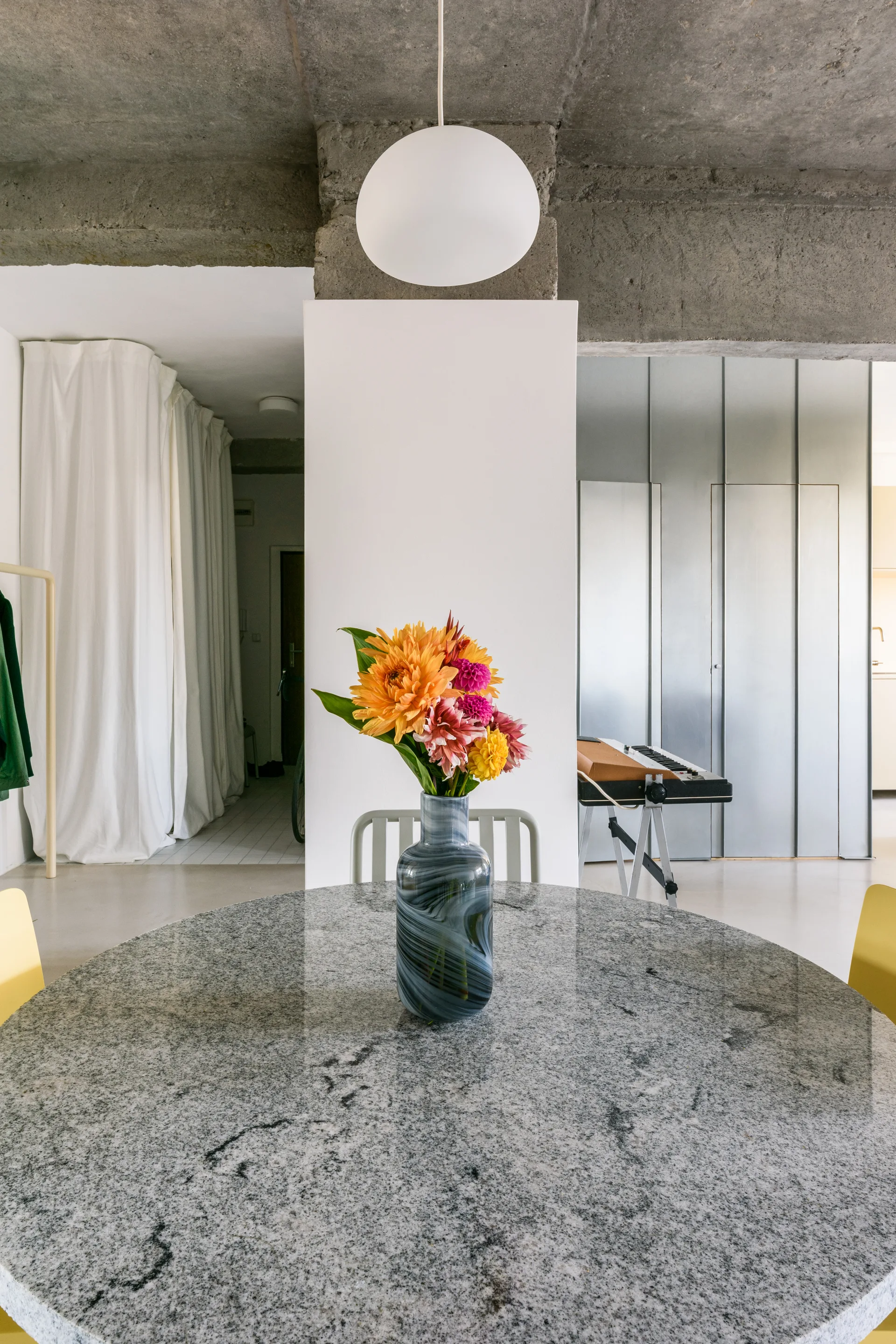 A round granite dining table holds a colorful floral arrangement under a white globe pendant, set against raw concrete and brushed metal walls.