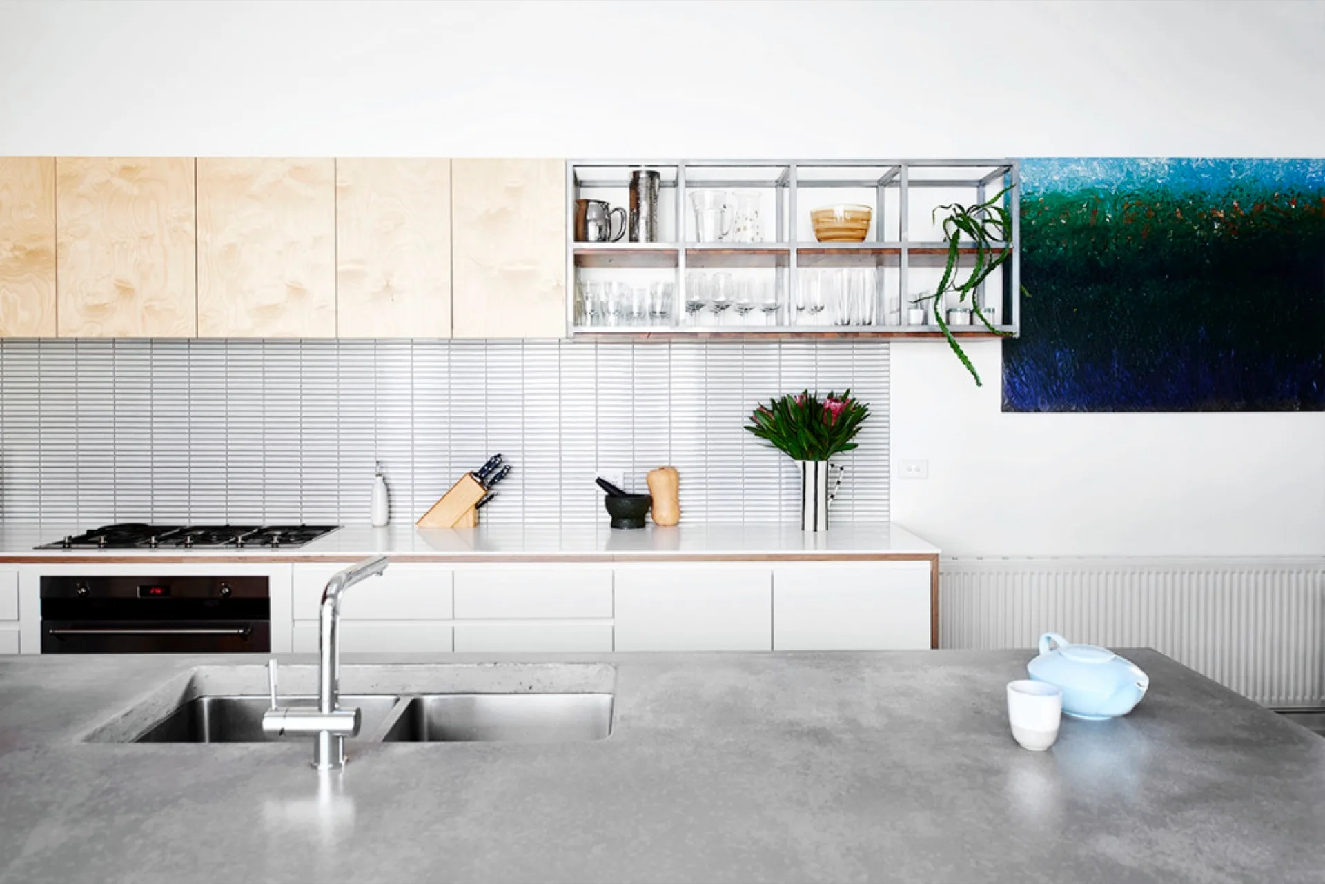 A modern kitchen features a grey concrete island with a double sink, light wood upper cabinets, and a white linear tile backsplash.