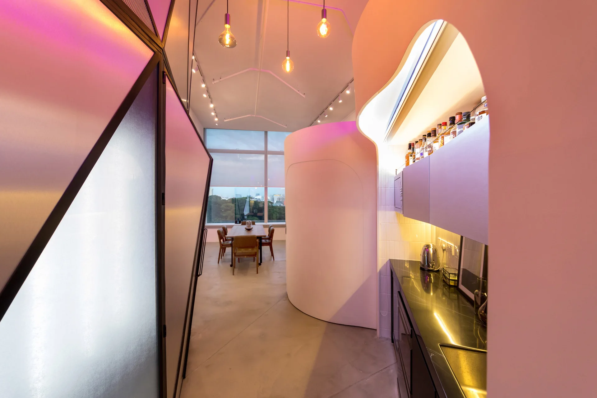 A modern interior corridor featuring a curved white partition wall, black kitchen countertop, hanging lightbulbs, and a dining table in the background.
