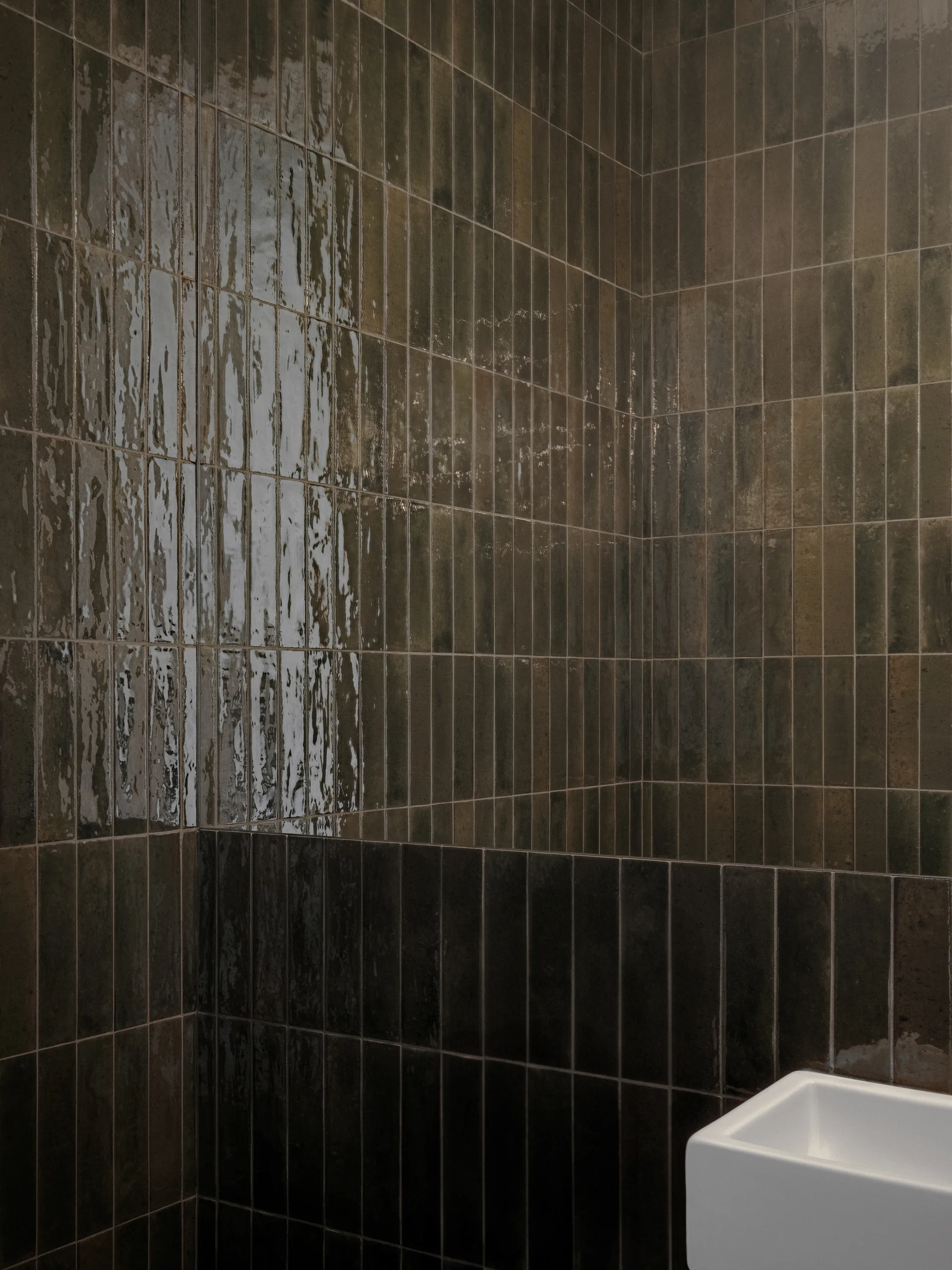 A close-up corner view of a bathroom featuring vertical stacked dark brown glossy wall tiles next to a white ceramic sink.