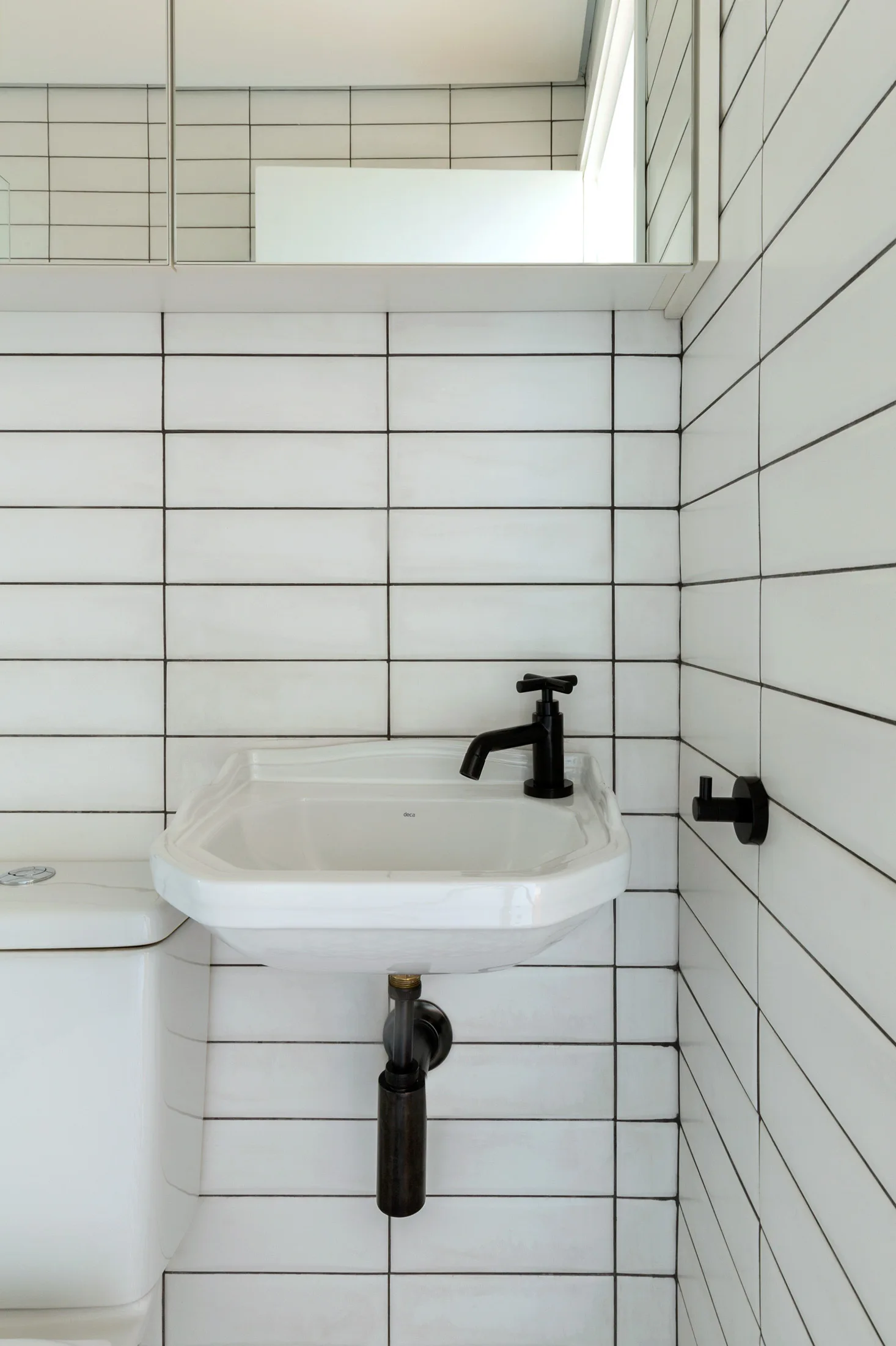 A small bathroom features a white wall-mounted sink with a matte black faucet and exposed plumbing against white rectangular tiles with dark grout.