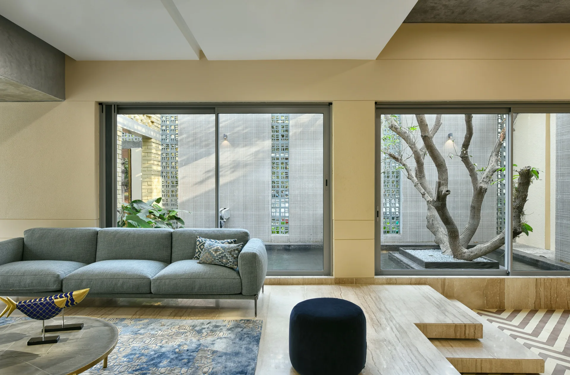 A modern living room features a grey sofa and blue rug next to large sliding glass doors that open to a courtyard with a tree.