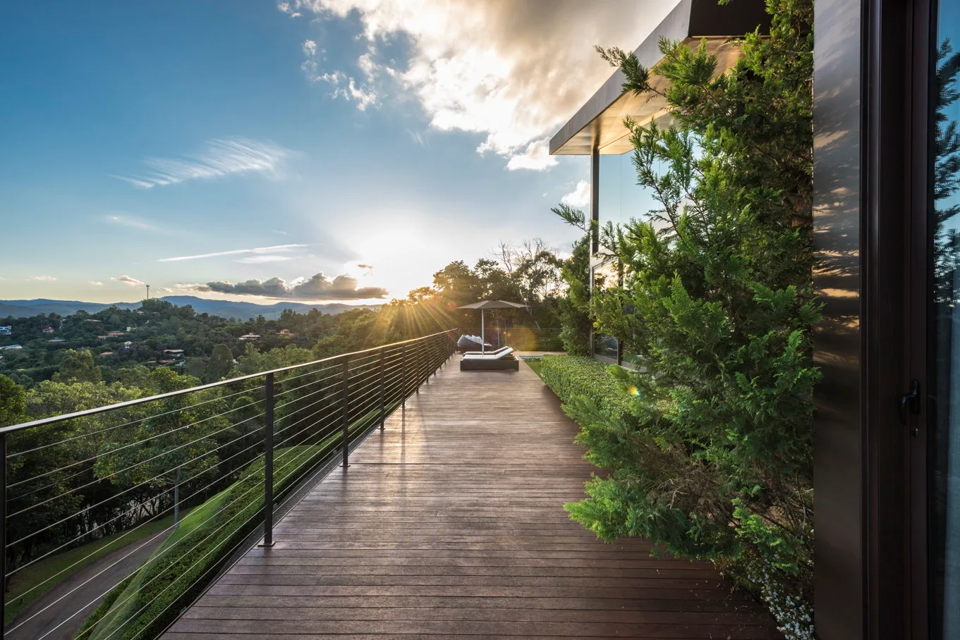 A long wooden terrace deck with a metal cable railing overlooks a green valley under a sunset sky.