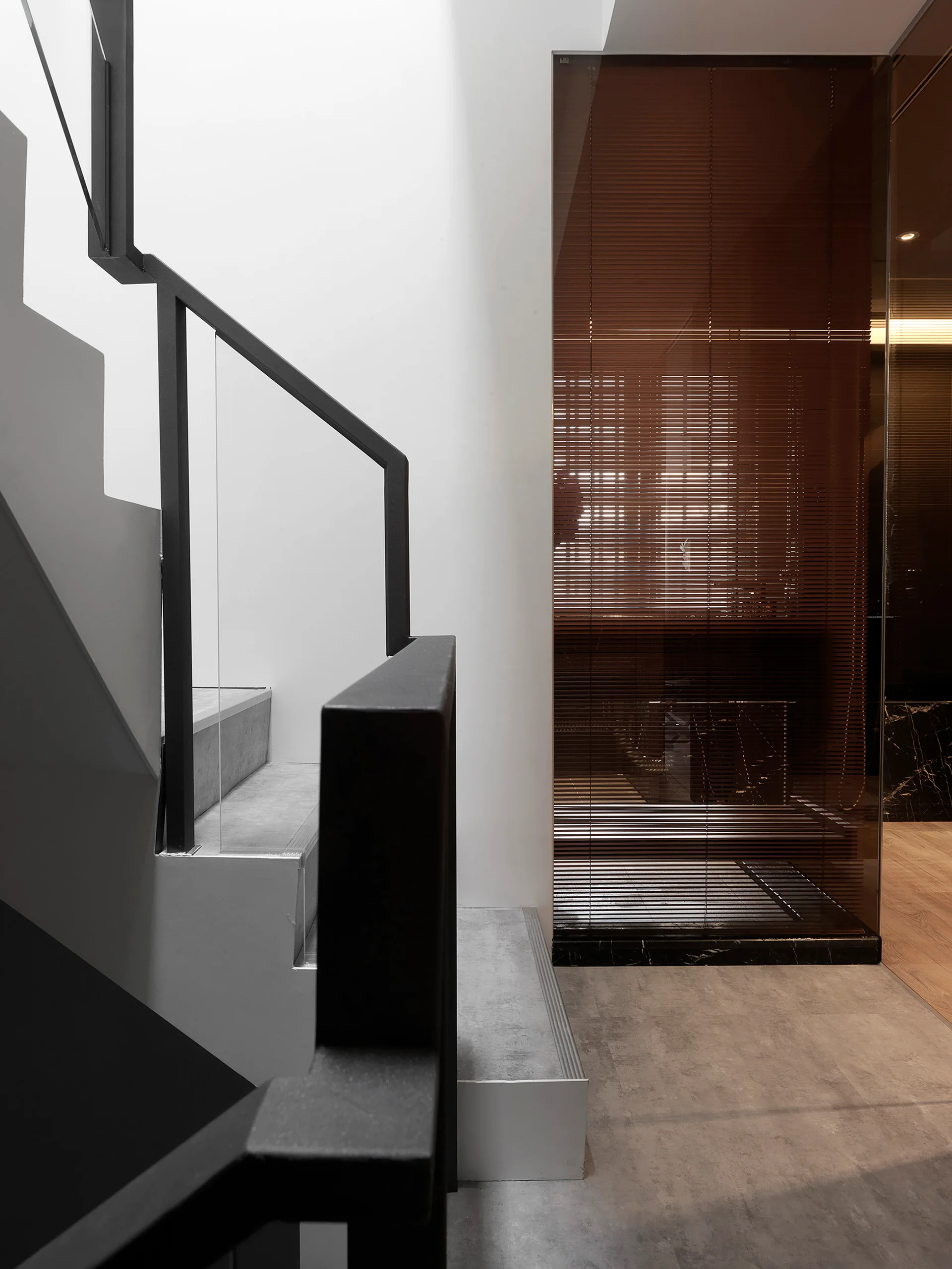 A modern staircase features gray concrete treads, a matte black metal railing with glass panels, and an adjacent glass wall enclosing brown blinds.