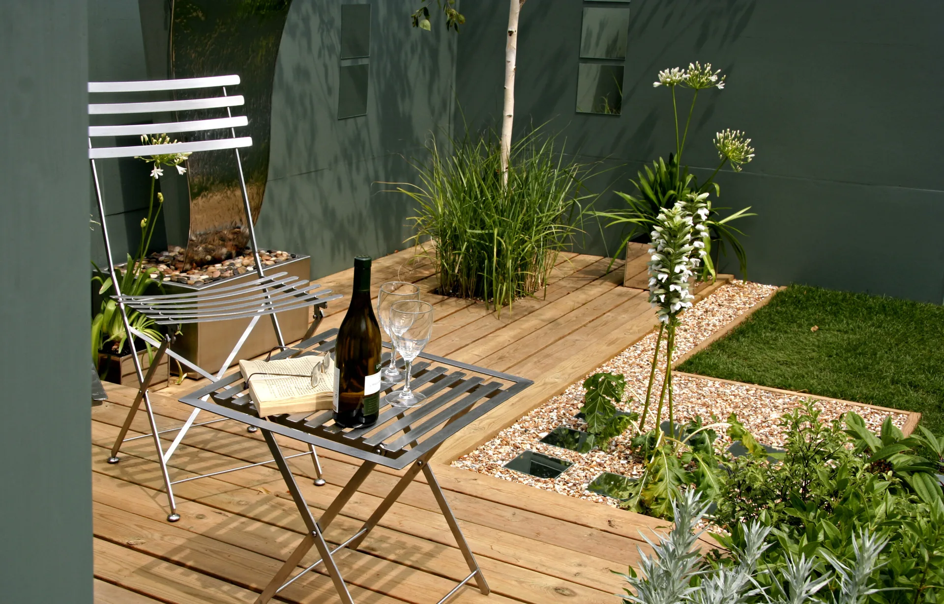 A metal bistro table and folding chair sit on a wooden deck surrounded by pebble-lined garden beds, a tall green wall, and decorative plants.