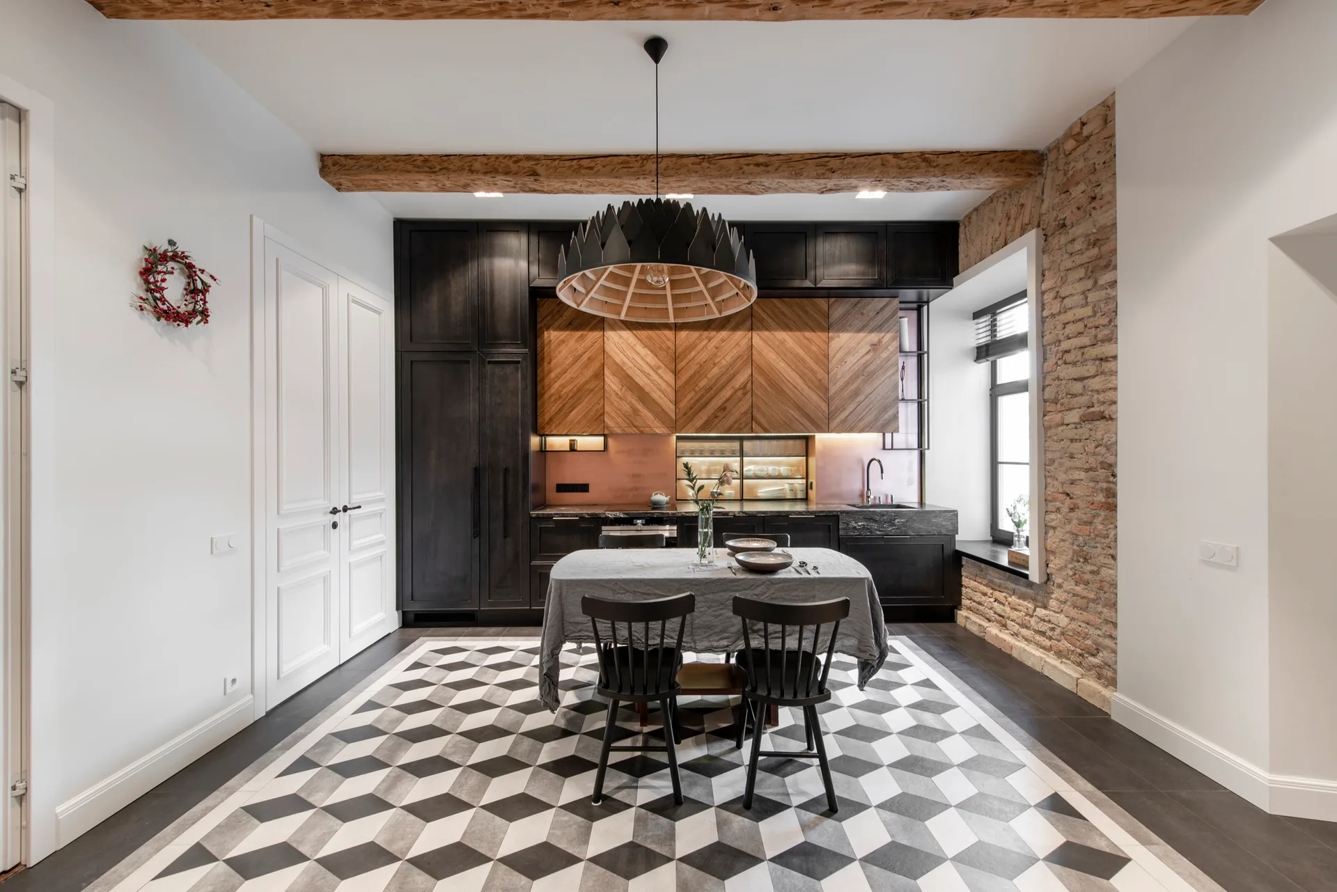A kitchen featuring black and herringbone wood cabinets, a geometric patterned tile floor, a rustic brick wall, and a dining table with two chairs.