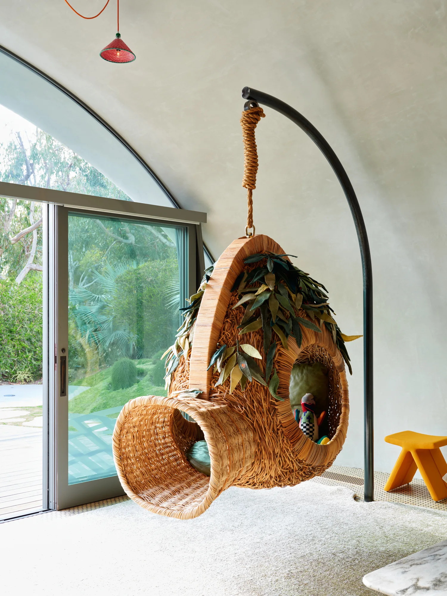A sculptural woven wicker hanging chair stands on a textured light floor next to a large sliding glass door with a view of a garden.