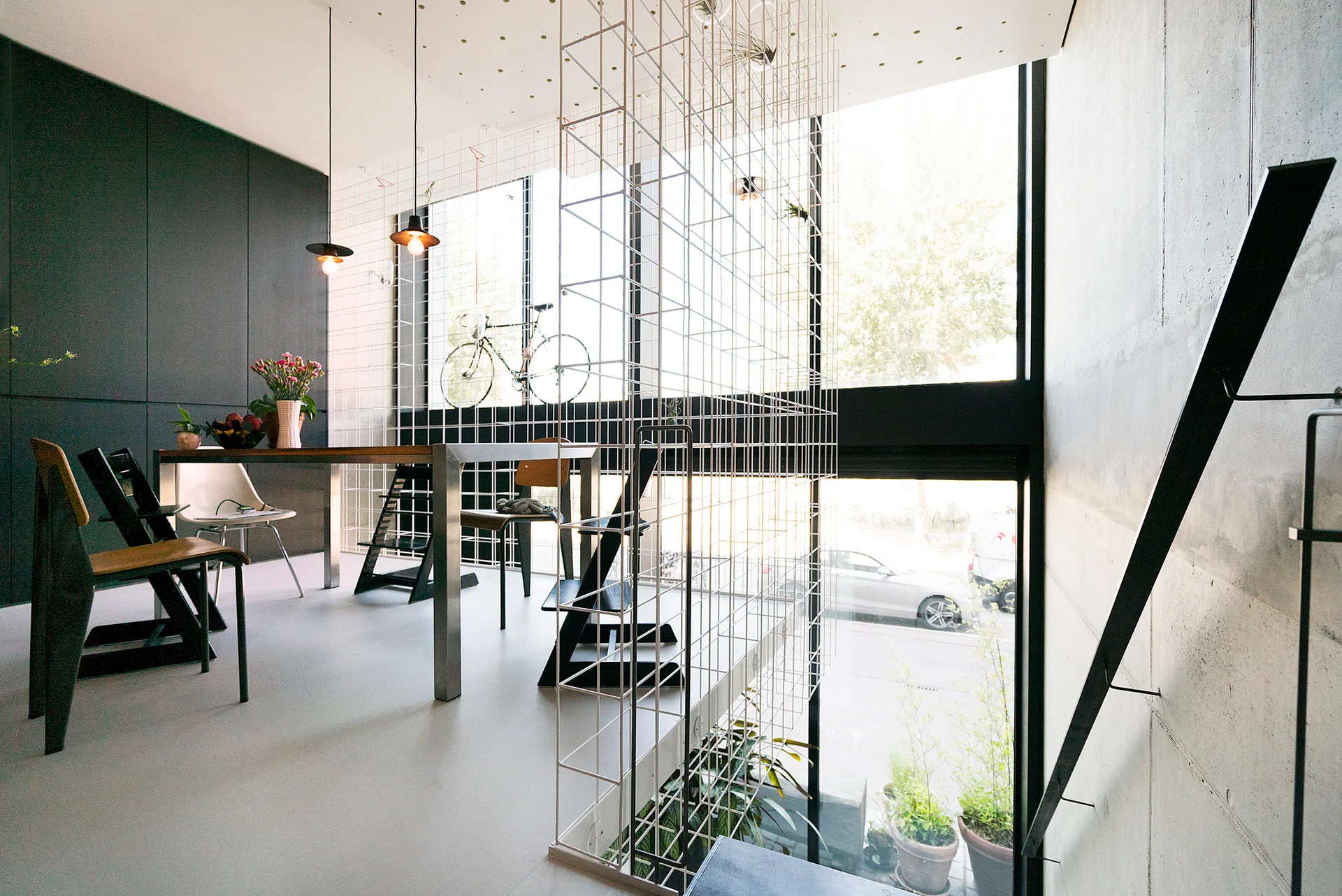 A minimalist dining area featuring a metal dining table, wood chairs, a tall white wire grid partition, and raw concrete walls next to a large window.